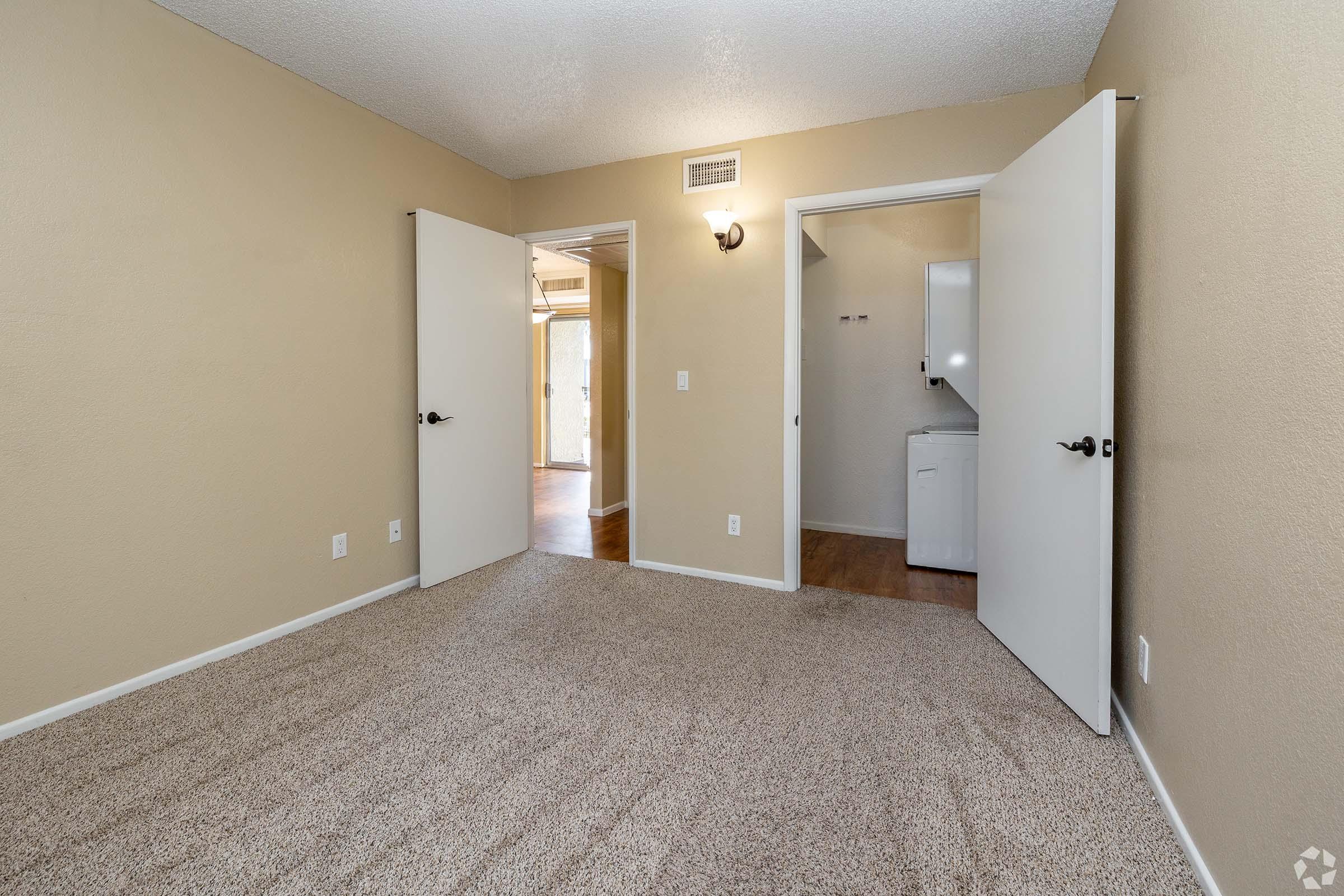 A spacious empty room with beige walls and light carpet. Two doors on the left lead to adjacent areas, and a doorway on the right opens to a laundry area. Natural light enters from the left side of the image, creating a bright and inviting atmosphere.