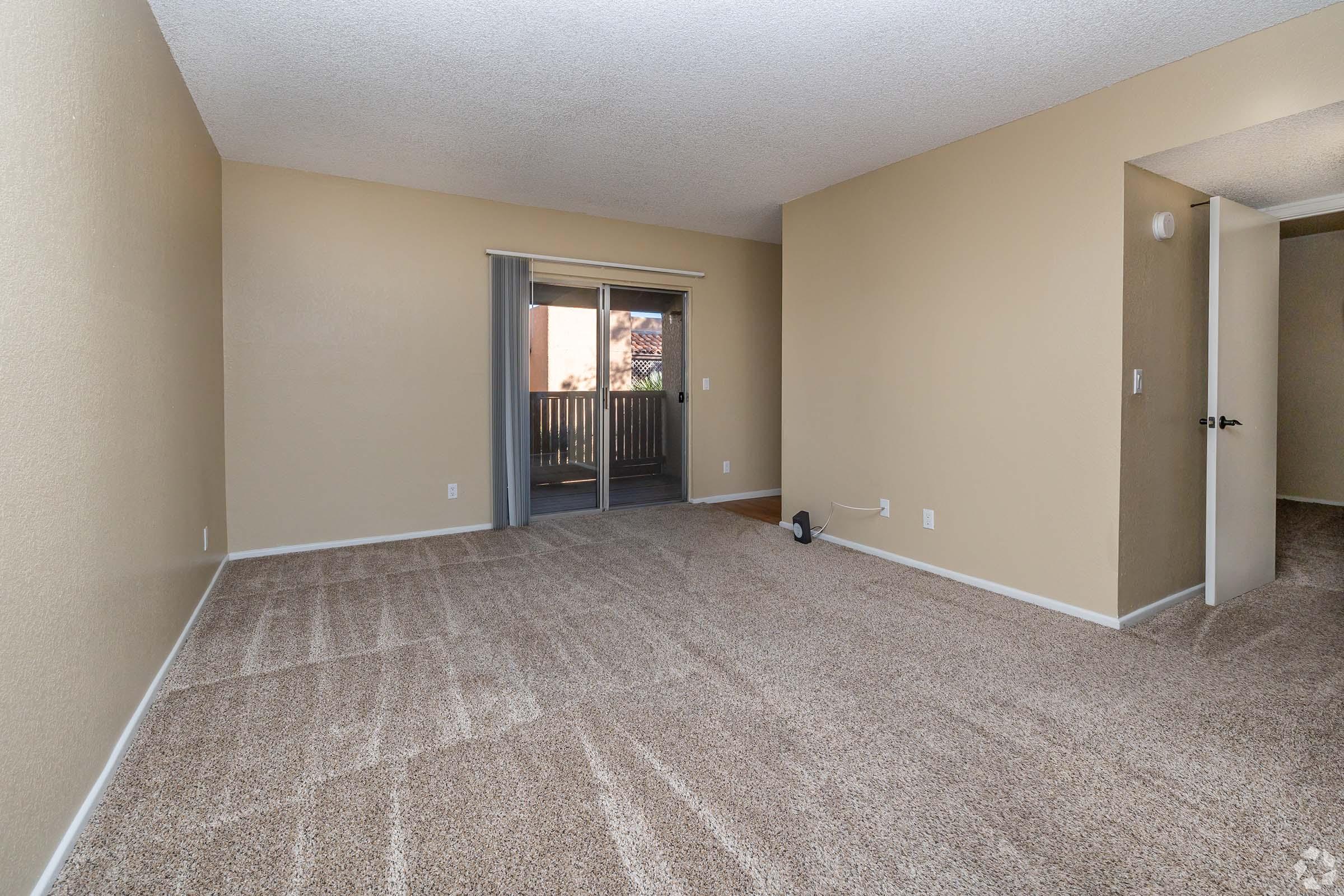 A spacious living room with light beige walls and carpet. There is a sliding glass door leading to a small balcony, with natural light entering the room. The room is empty, featuring a closet door on the right and an overall clean, minimalistic design.