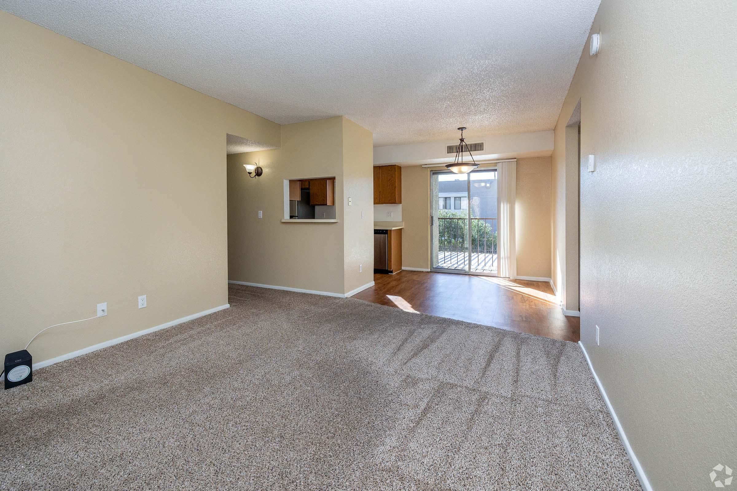 A well-lit living room with beige walls and carpet, featuring a doorway leading to a kitchen area with wooden cabinets. There is a sliding glass door that opens to an outdoor space, with natural light casting shadows on the floor. The room is minimally decorated and spacious, creating a cozy atmosphere.