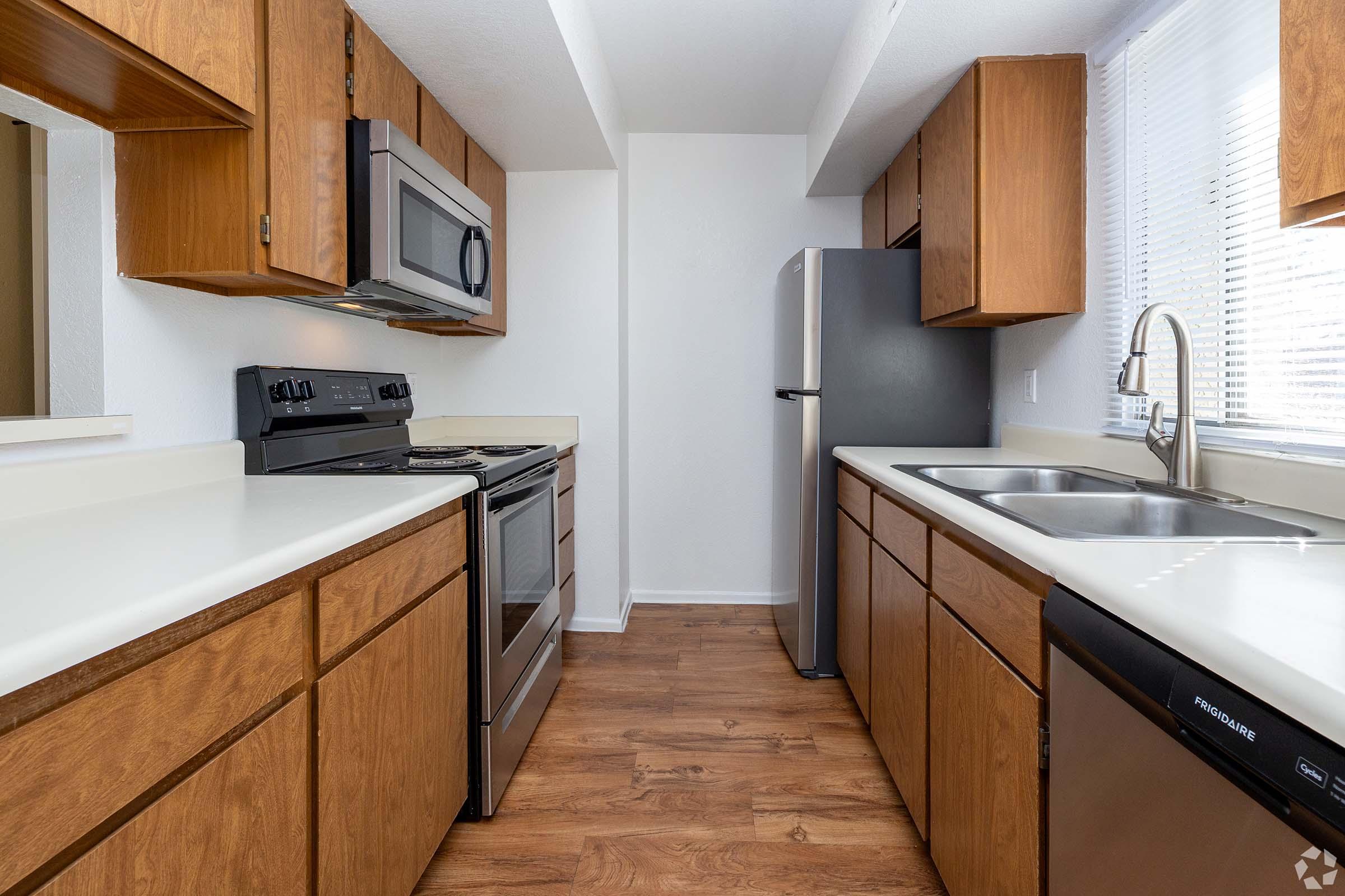 A modern kitchen with wooden cabinets, stainless steel appliances, including a microwave, oven, and refrigerator. The countertops are light-colored, and there's a dual sink by a window. The flooring is a warm wood tone, creating a bright and inviting atmosphere.