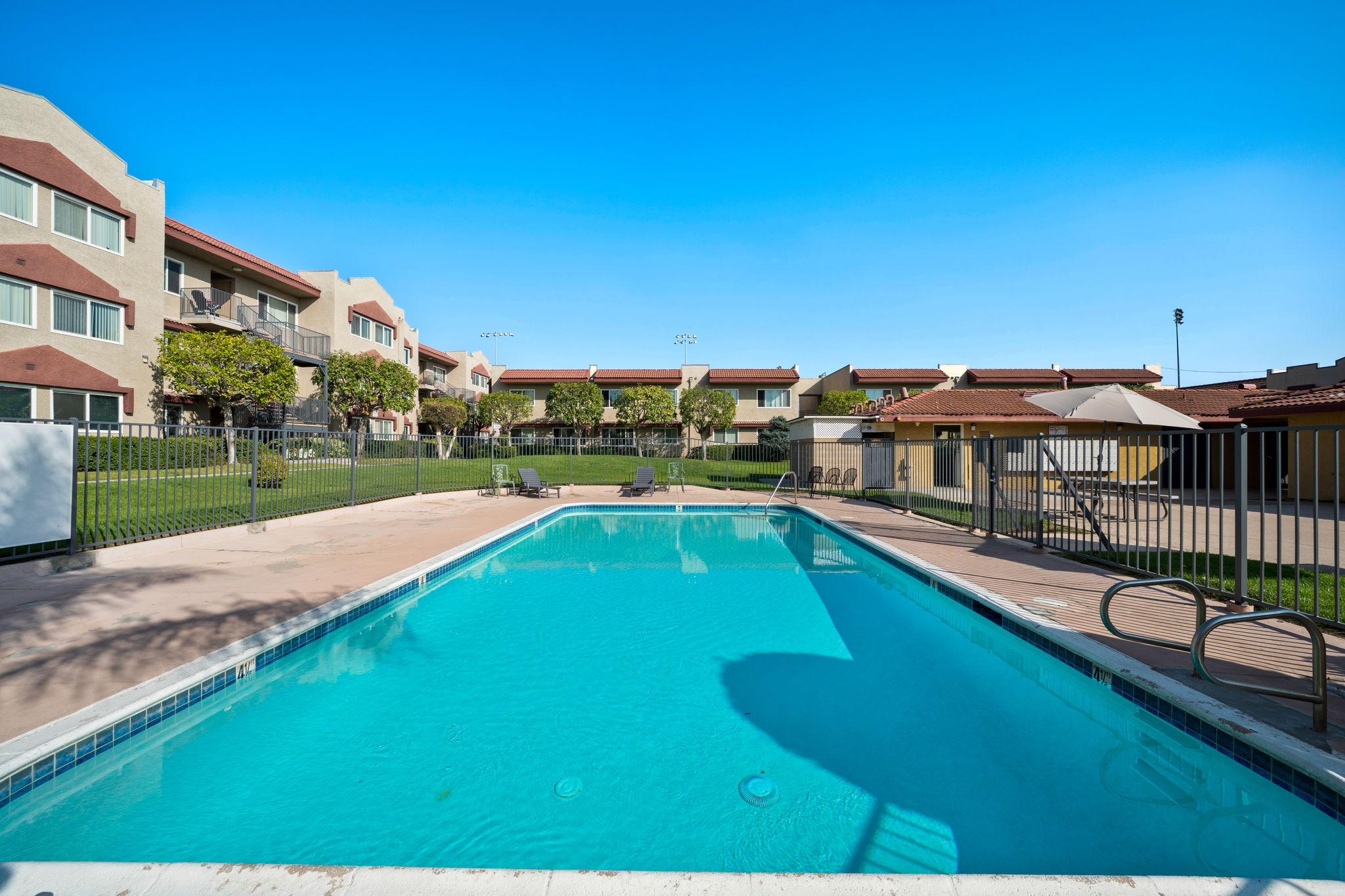 A clear blue swimming pool surrounded by a fence, with lounge chairs on the pool deck. In the background, there are two-story residential buildings and well-maintained greenery under a bright blue sky. The scene conveys a peaceful, sunny day perfect for relaxation.