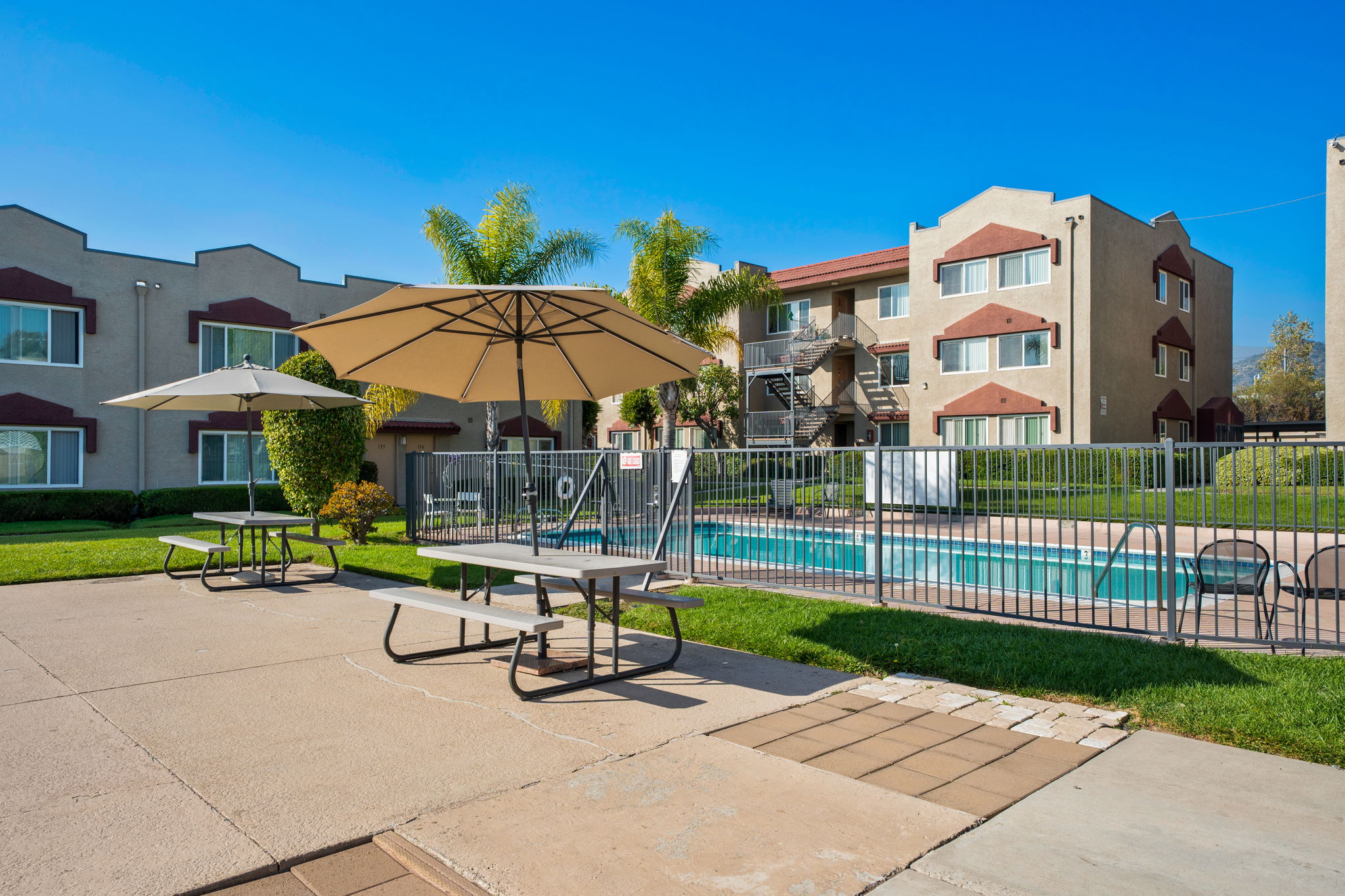 A sunny view of a residential complex featuring a swimming pool, surrounded by green grass and palm trees. There are picnic tables with umbrellas, and the building has multiple stories with colorful accents. The clear blue sky complements the inviting atmosphere of the space.