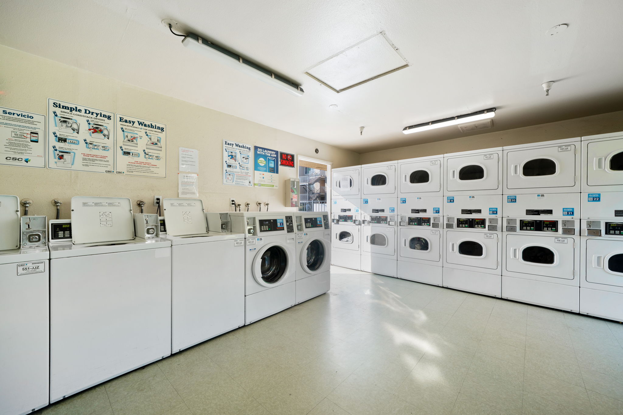 A well-lit laundry room featuring multiple white washing machines and dryers lined up against the walls. The space includes informational signs on the walls about laundry services. The floor is light-colored, and there are fluorescent lights on the ceiling, creating a clean and tidy environment.