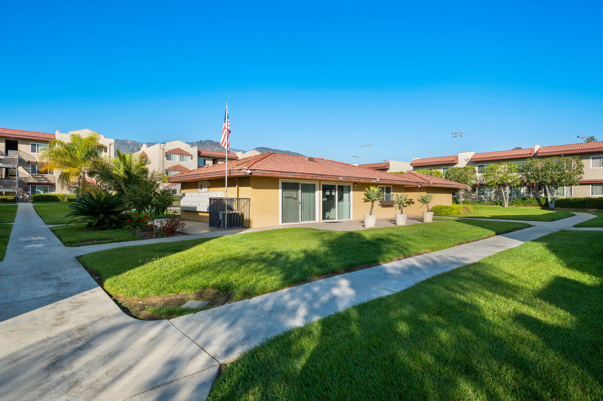 A well-maintained residential complex with a yellow building, greenery, and palm trees. The building features a red tile roof and large windows. There are curved pathways and grassy areas leading to the entrance, with a clear blue sky above and mountains visible in the background.