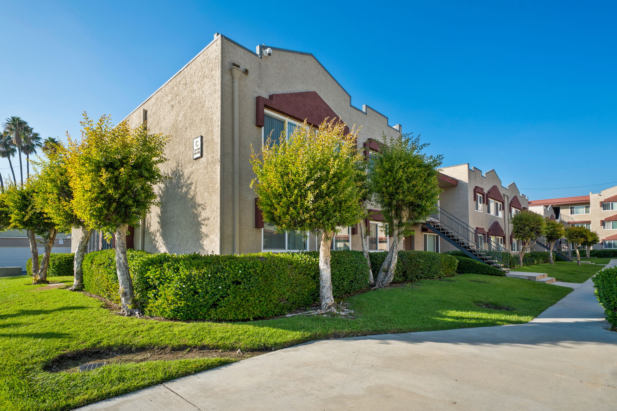 A well-maintained two-story apartment building with a light-colored exterior, surrounded by manicured shrubs and small trees. A clear blue sky is above, and a walkable path leads to the entrance. The complex features a modern design with stairs leading to upper units.