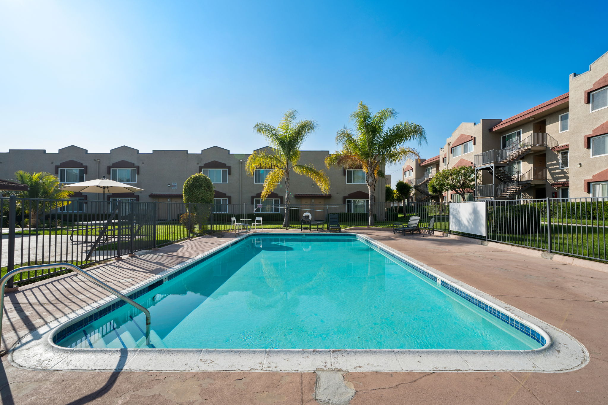 A clear blue swimming pool surrounded by a relaxing patio area, featuring lounge chairs and umbrellas. Lush green palm trees provide a tropical atmosphere, while the backdrop includes modern apartment buildings under a bright blue sky. Perfect for leisure and relaxation.