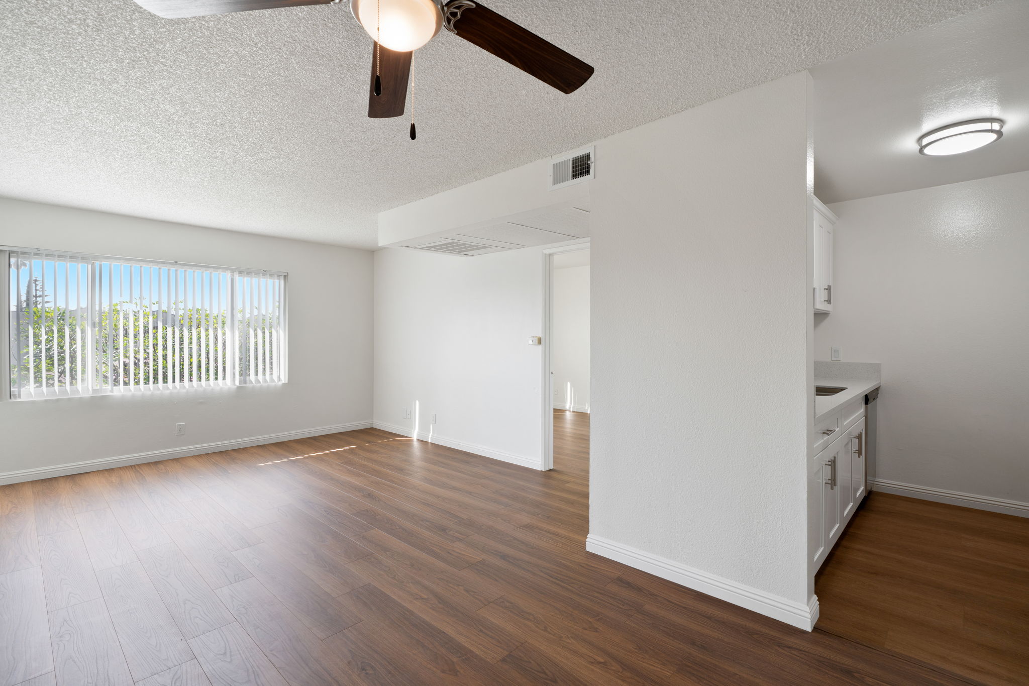 A bright, spacious room with wood-like flooring and a ceiling fan. The room features large windows with blinds, allowing natural light in. A modern kitchenette is visible on the right, with white cabinets and countertops. The walls are painted white, creating a clean and airy atmosphere.