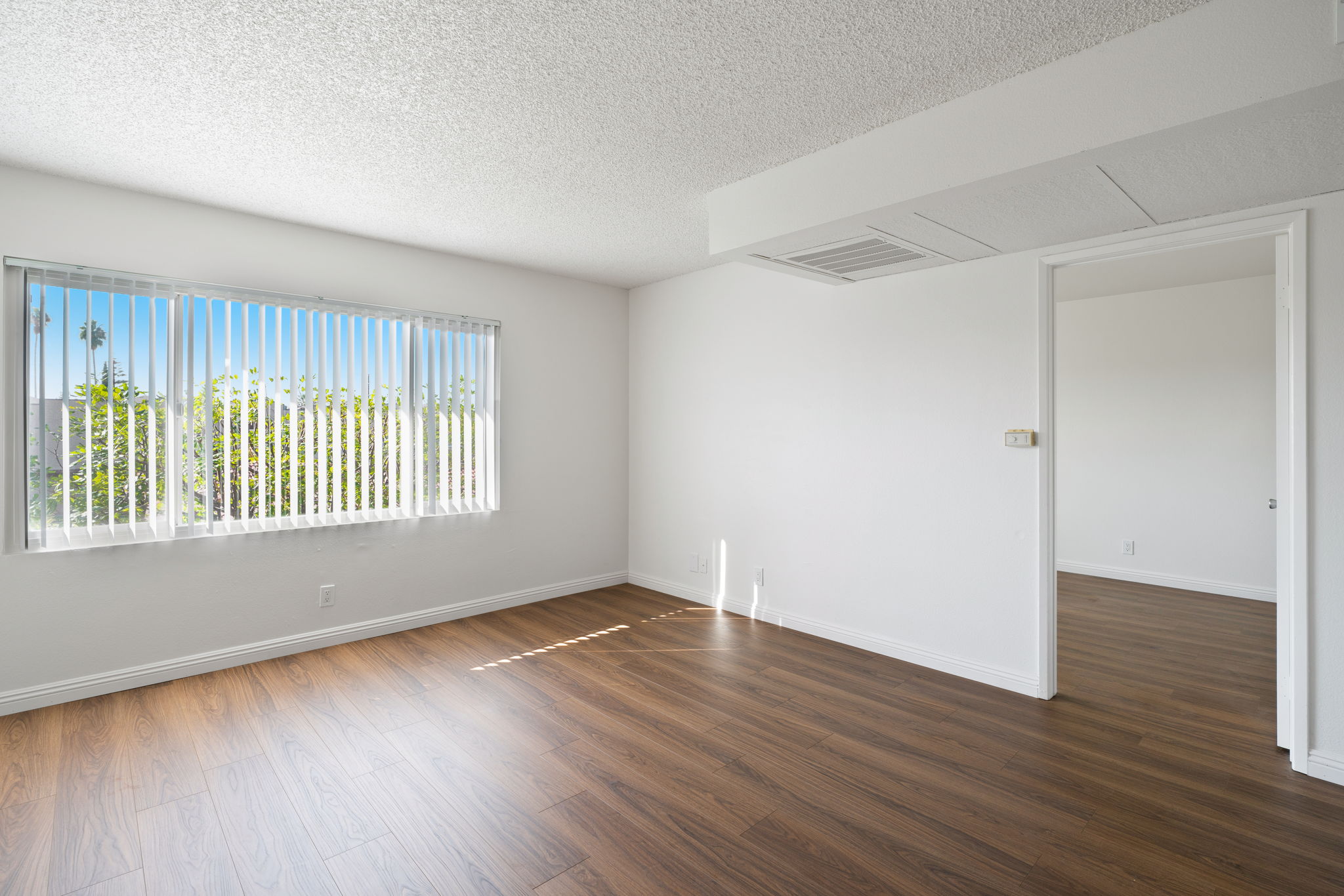 Bright and spacious empty room with light-colored walls and a large window featuring vertical blinds. The floor is wood laminate, and there is a small vent on the ceiling. In the background, another doorway leads to an adjacent room. Natural light fills the space, creating an inviting atmosphere.