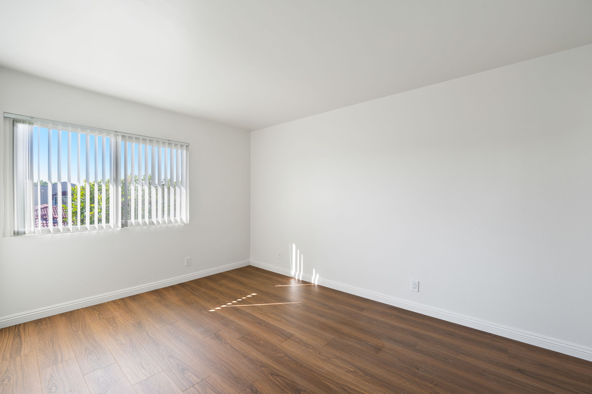 Bright, empty room featuring light-colored walls and wooden flooring. A window with vertical blinds allows natural light to enter, illuminating the space. The room has a minimalistic design with no furniture, creating a spacious and clean atmosphere.