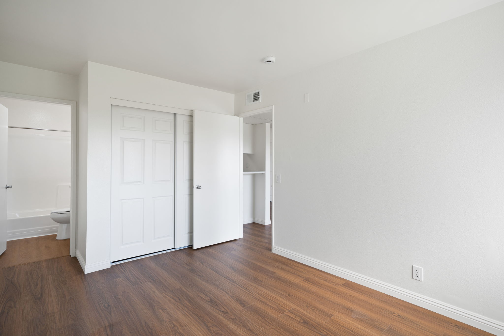 A spacious, empty room featuring light-colored walls and wooden flooring. There are sliding closet doors on one wall and a doorway leading to a bathroom visible in the background. The overall ambiance is bright and clean, suitable for various uses such as a bedroom or office.