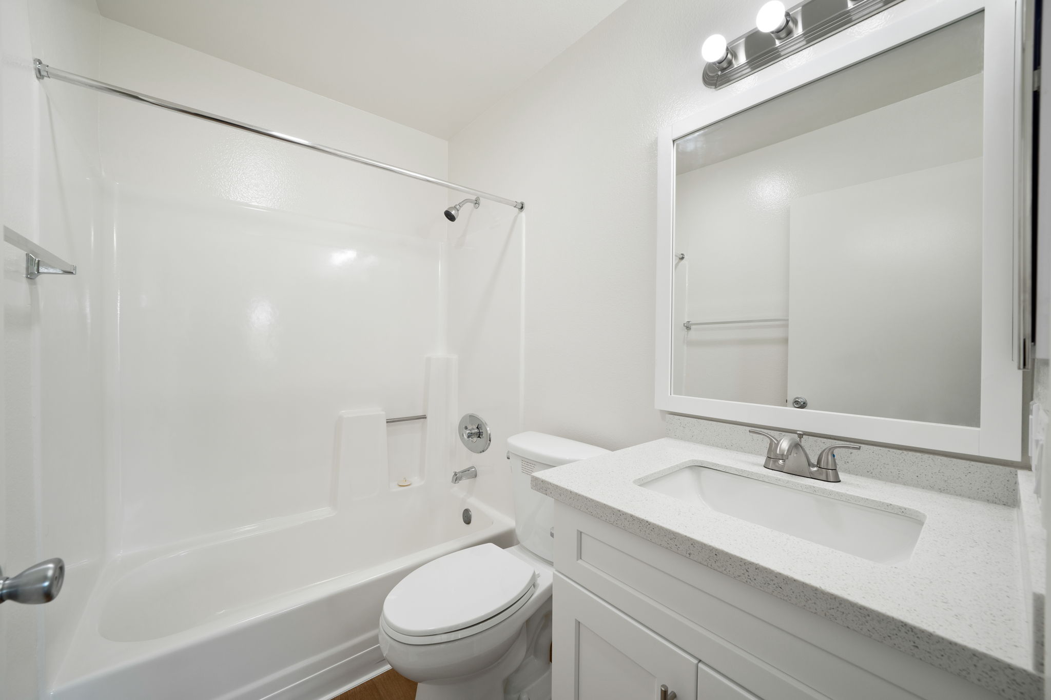 A clean and modern bathroom featuring a bathtub with a shower, a white sink with a mirror above, and a toilet. The walls are painted white, and the room is well-lit with overhead lighting. The floor is made of light-colored wood.