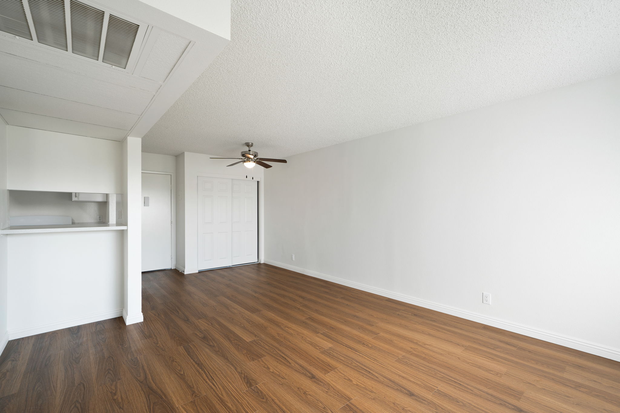 A spacious, well-lit room with light brown laminate flooring, featuring a ceiling fan and white walls. There is an open area leading to a kitchen with a bar counter. A closet door is visible on the left, and on the right, a white door leads to another room. The overall look is clean and modern.