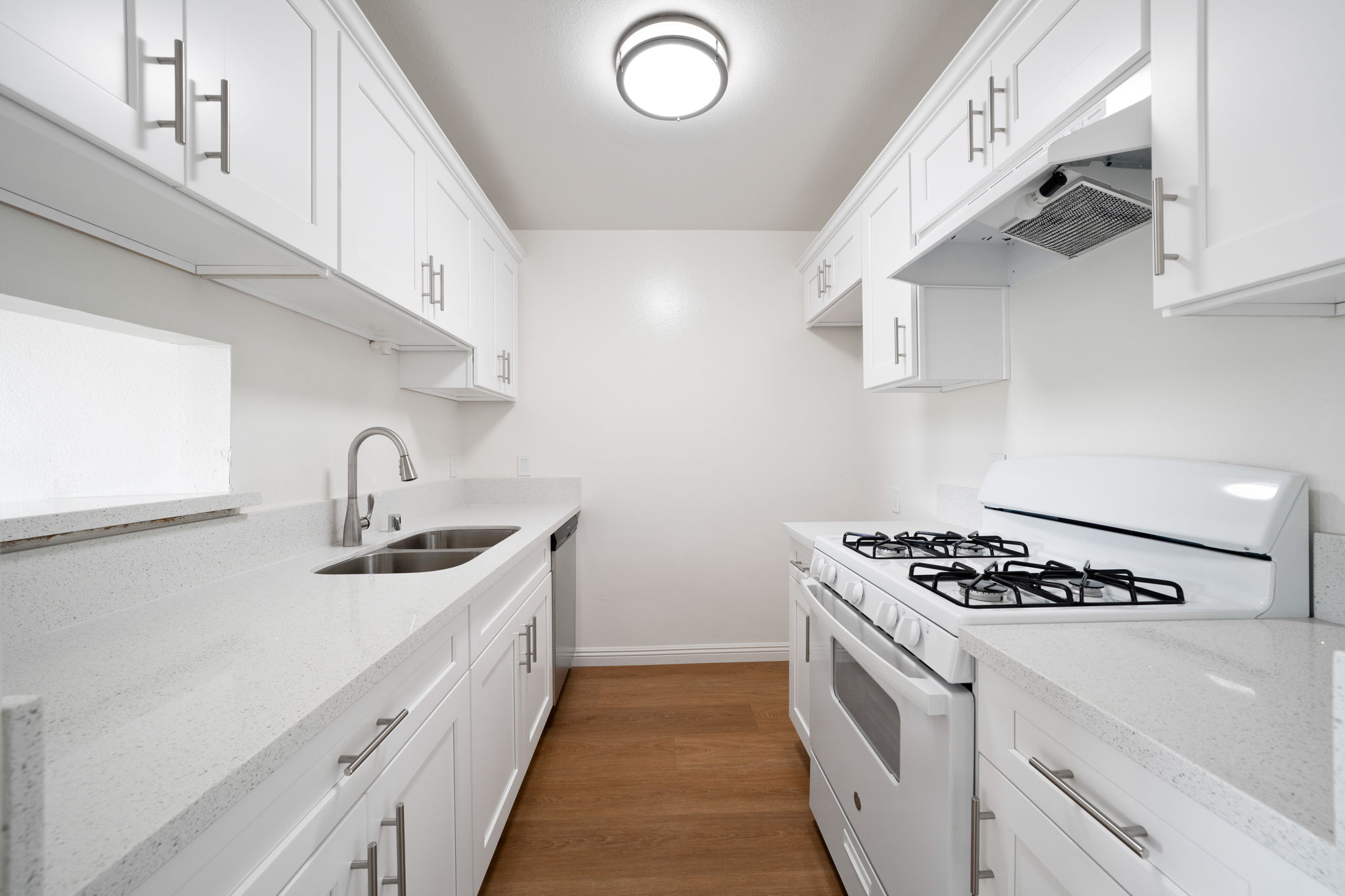 A modern kitchen featuring white cabinets, a double sink, and a gas stove. The countertops are light-colored, complementing the cabinetry. A ceiling light fixture provides illumination, and the space has a clean, minimalist design with wooden flooring.