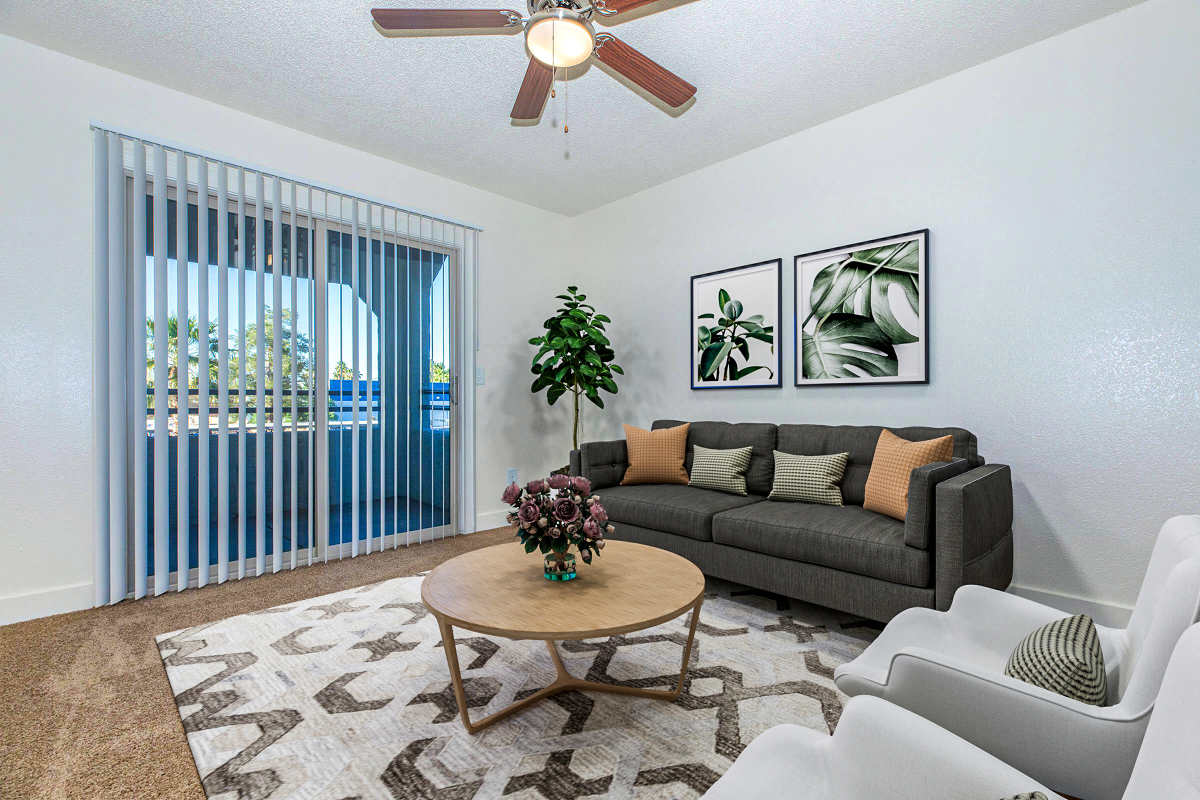 A cozy living room featuring a gray sofa with decorative cushions, two white armchairs, a round wooden coffee table with a floral arrangement, and a decorative rug. Large sliding glass doors with vertical blinds open to a balcony. The walls are adorned with framed botanical art and a potted plant adds a touch of greenery.