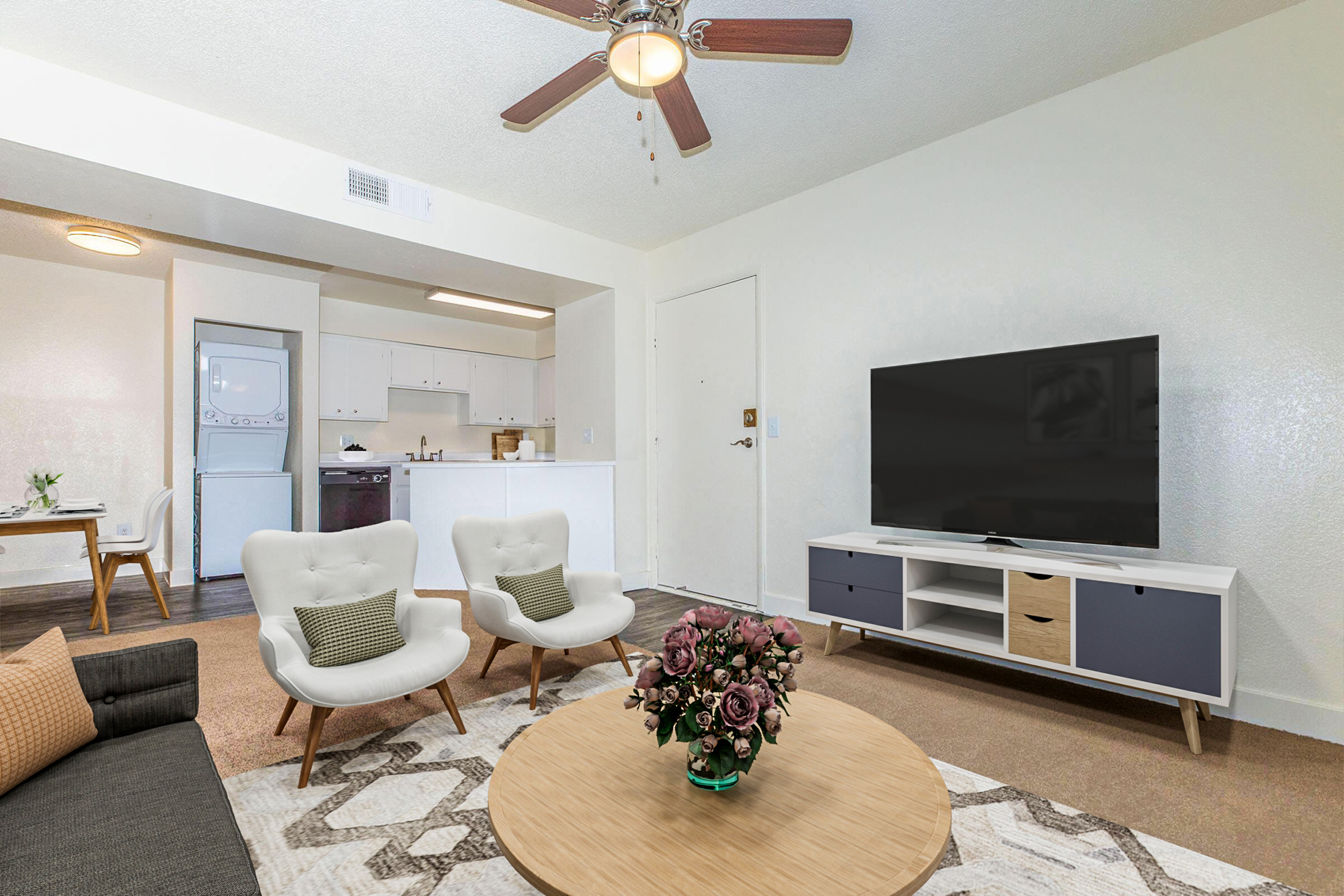 A modern living room featuring a neutral color palette. There are two white armchairs with green cushions facing a wooden coffee table with a floral arrangement. A large flat-screen TV is on a white TV stand against the wall. The open kitchen can be seen in the background, with a dining area included.