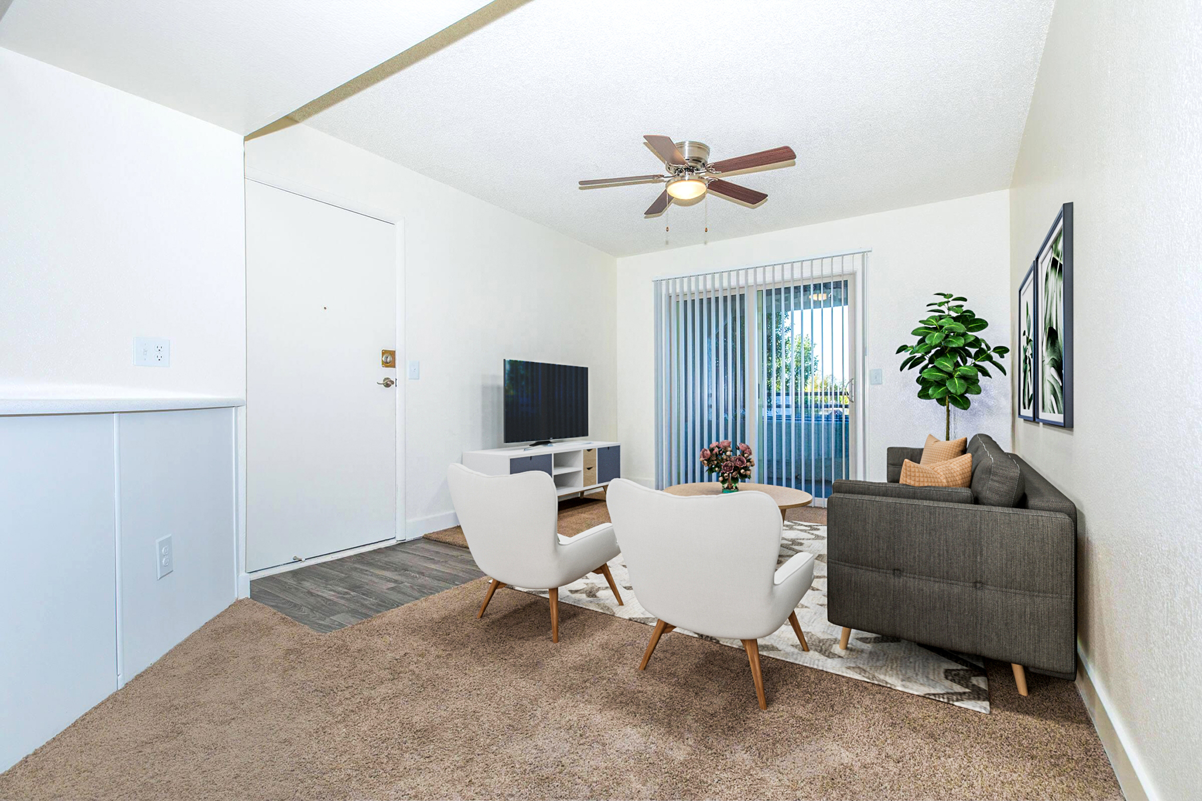 A cozy living room featuring two stylish chairs, a gray sofa, a TV on a white stand, and a decorative plant. Light-colored walls and a ceiling fan add to the ambiance, while large windows with vertical blinds let in natural light. A small table with flowers completes the inviting space.