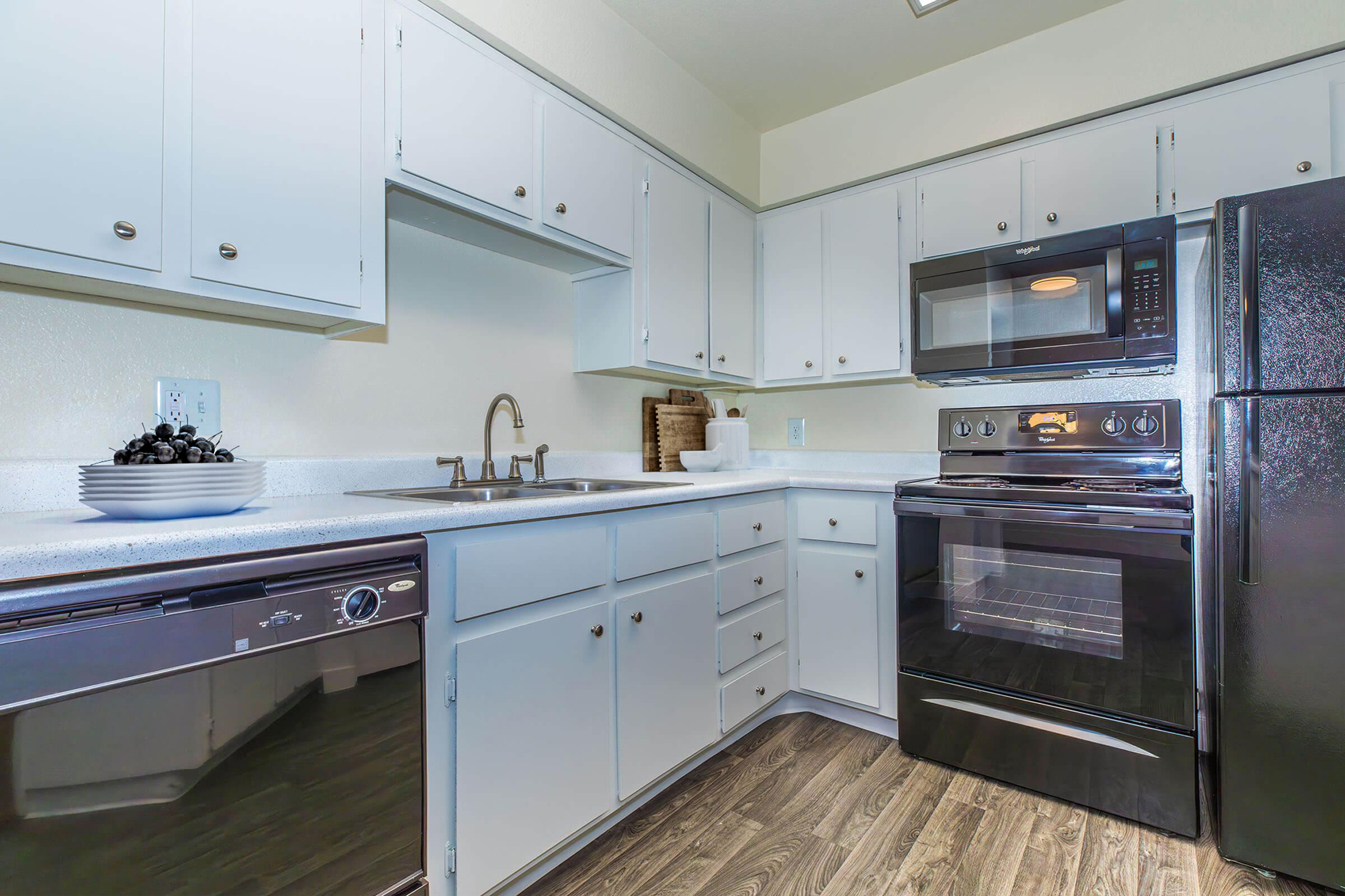 A modern kitchen featuring white cabinets, a stainless steel sink, an electric stove, a microwave, and a black refrigerator. The countertop is clean and organized, with a bowl of decorative items on it. The flooring is wood-like, creating a warm and inviting atmosphere.