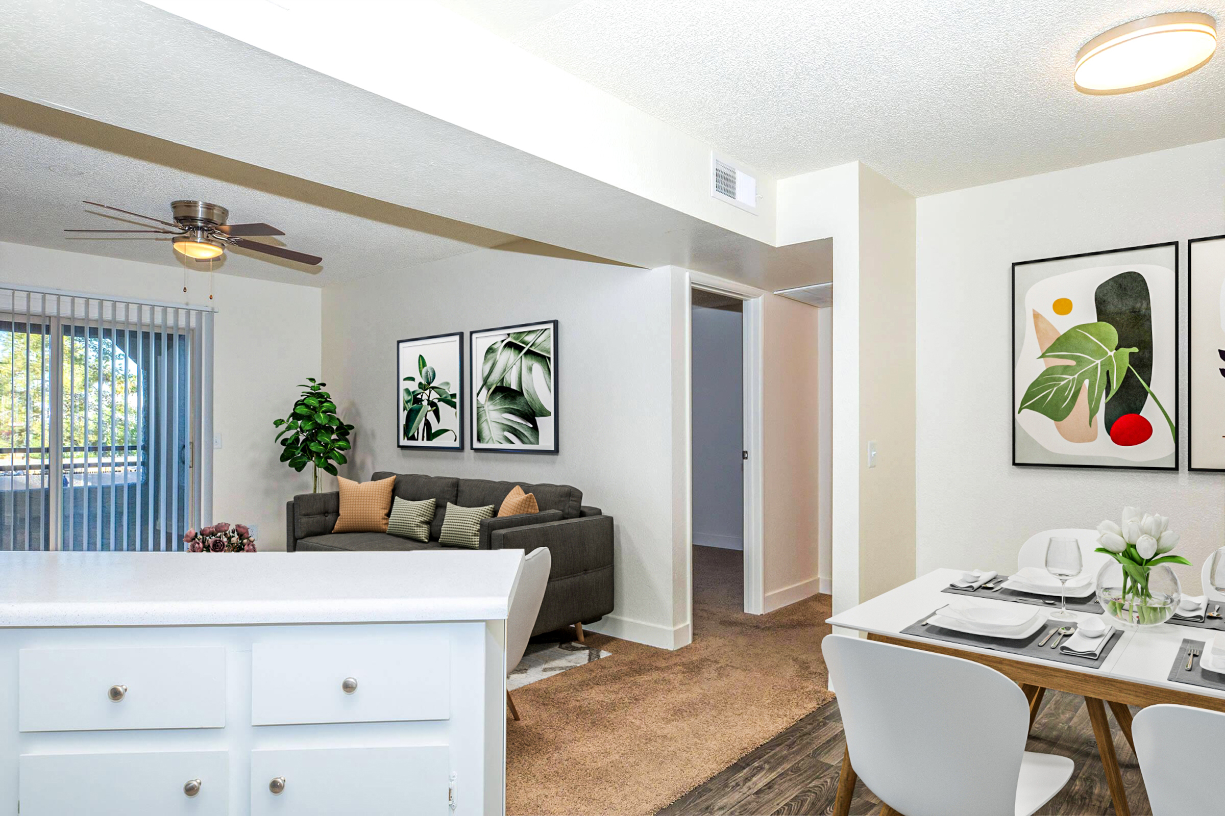 A modern living area featuring a comfortable grey sofa, a dining table set for two, and decorative artwork on the walls. A potted plant adds a touch of nature, while sliding glass doors provide natural light. The space has a clean, inviting aesthetic with neutral tones and a ceiling fan.
