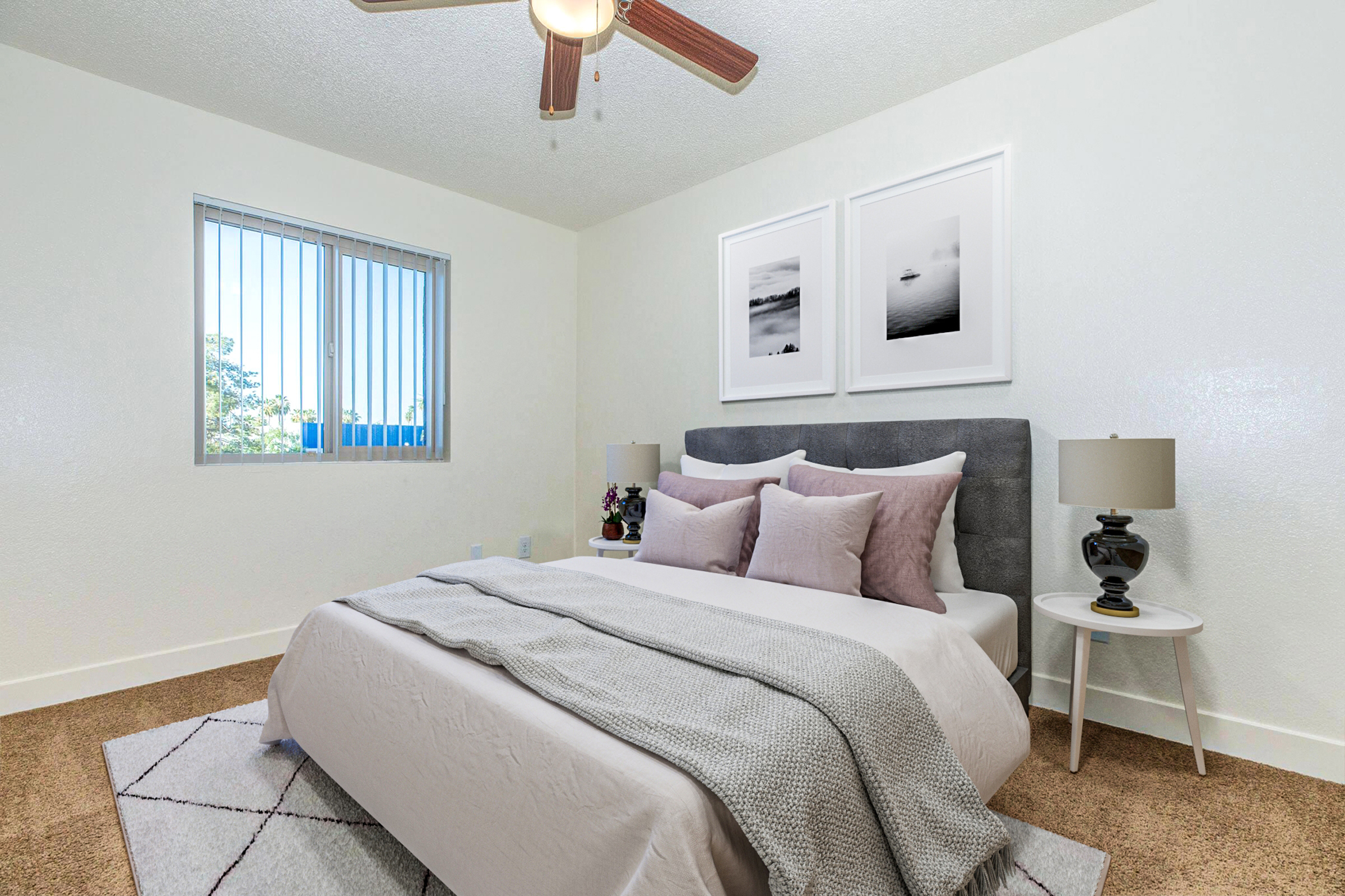 A bright and cozy bedroom featuring a large bed with soft pillows, a stylish gray headboard, and a light gray blanket. There are two decorative wall art pieces above the bed, and the room includes a ceiling fan and two bedside lamps on small tables. Natural light comes through a window with vertical blinds.