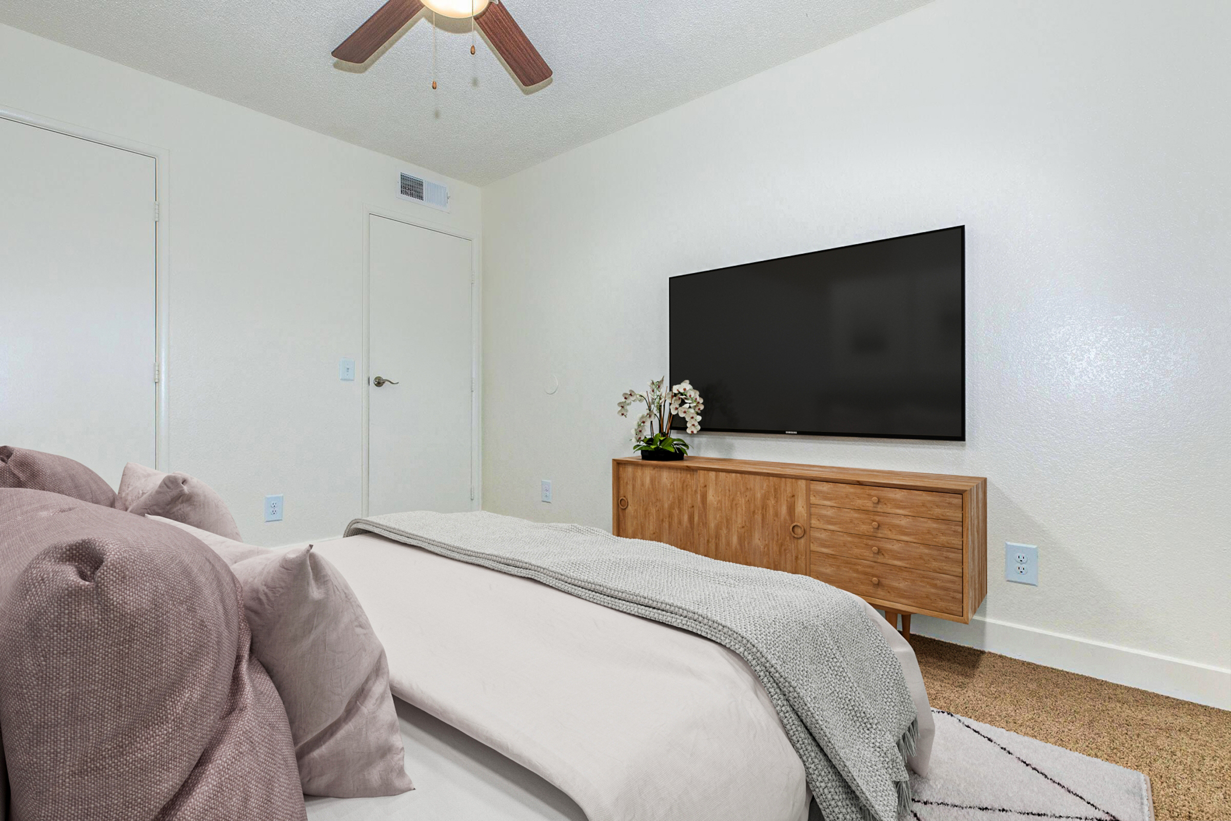 A cozy bedroom featuring a bed with soft, layered bedding, a wooden nightstand, and a modern TV mounted on the wall above a low-profile wooden cabinet. A ceiling fan with wooden blades adds charm, while a few decorative plants enhance the inviting atmosphere of the space. Bright, neutral walls create a calming feel.