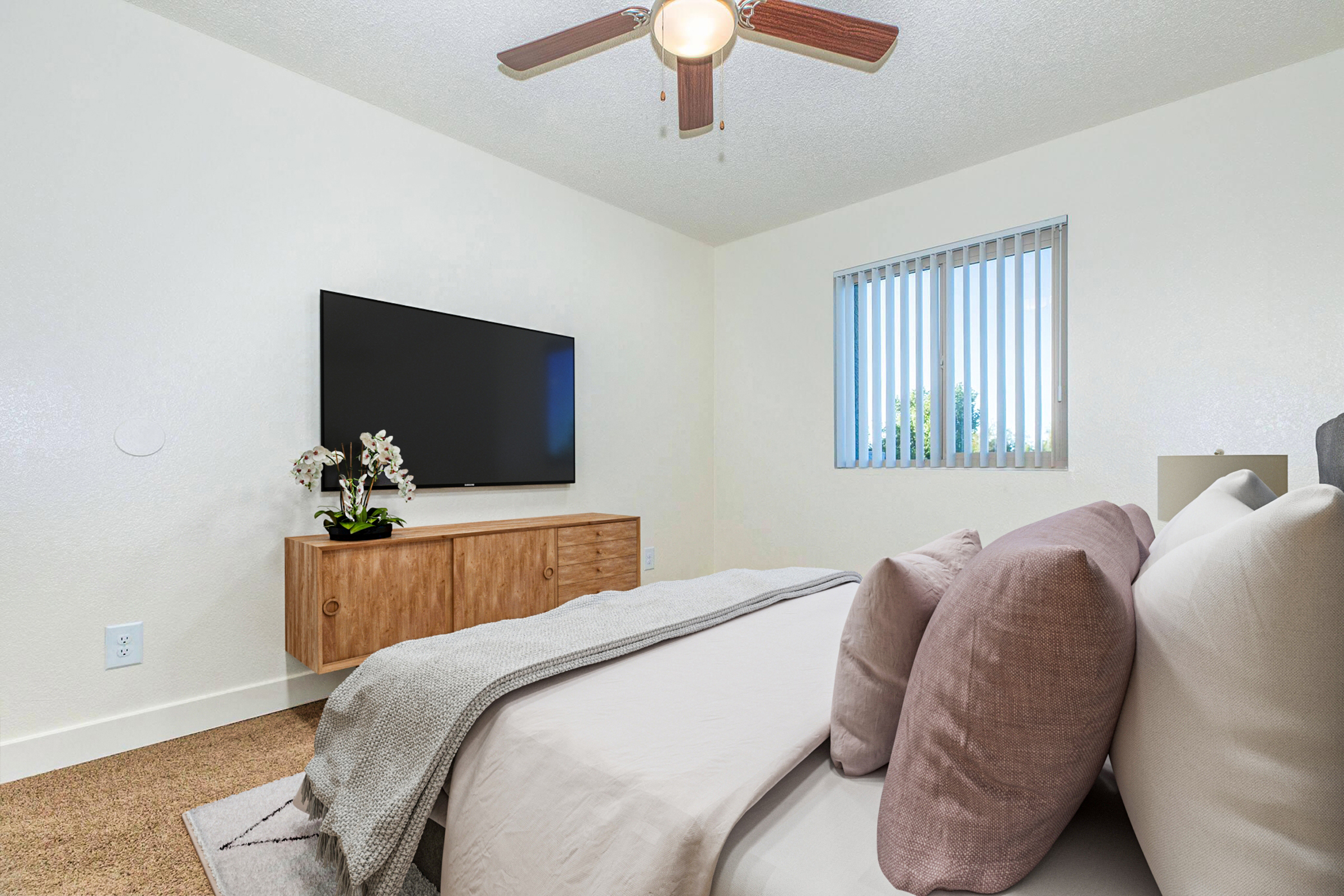 A neatly arranged bedroom featuring a bed with decorative pillows, a wooden dresser underneath a wall-mounted TV, and a window with vertical blinds. A potted orchid adds a touch of greenery, while a ceiling fan hangs overhead, enhancing the cozy atmosphere. The walls are painted a light color, contributing to a bright feel.