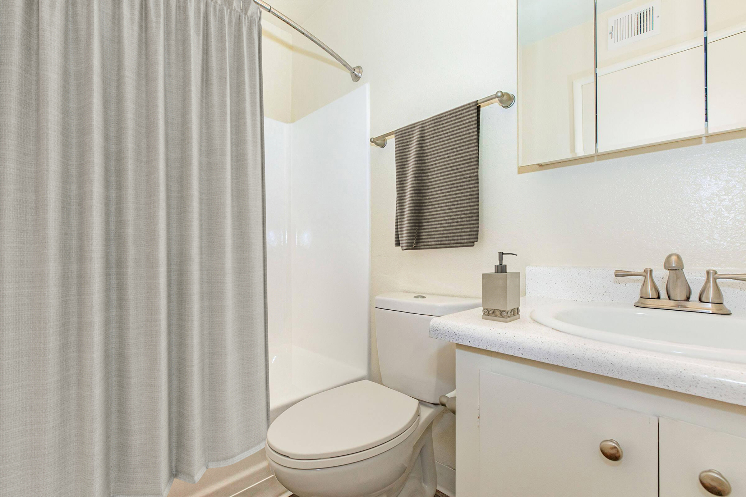 A modern bathroom featuring a white shower enclosure with a gray shower curtain, a toilet, and a sink with a countertop. There is a mirror above the sink and a gray towel hanging on a towel rack. The walls are painted light, creating a bright and clean aesthetic.
