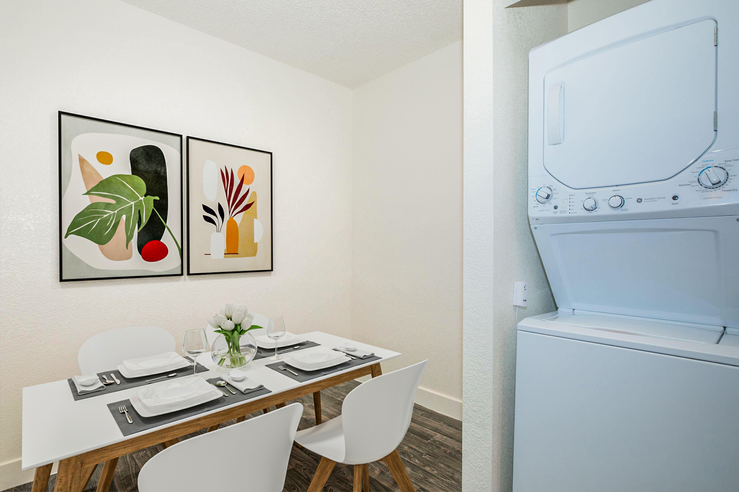 A modern dining area featuring a white table set for four with plates, cutlery, and a small vase of flowers. On the wall, two abstract art pieces with vibrant colors. To the right, a stacked washer and dryer in a compact space, adding convenience to the room. The overall aesthetic is clean and contemporary.