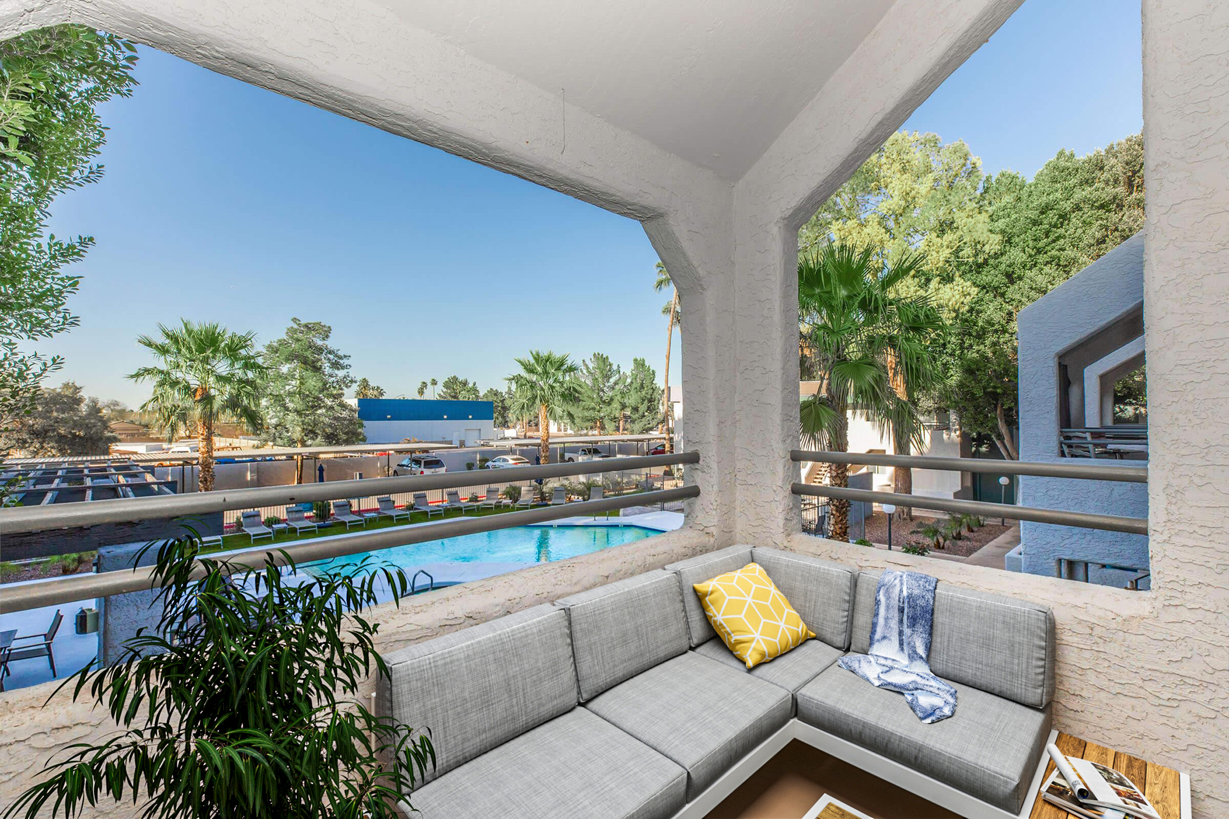 A cozy outdoor seating area with a gray sectional sofa adorned with a yellow decorative pillow. The view through a large open window features palm trees and a swimming pool in a sunlit courtyard. The surrounding architecture is modern and inviting, creating a relaxing atmosphere.