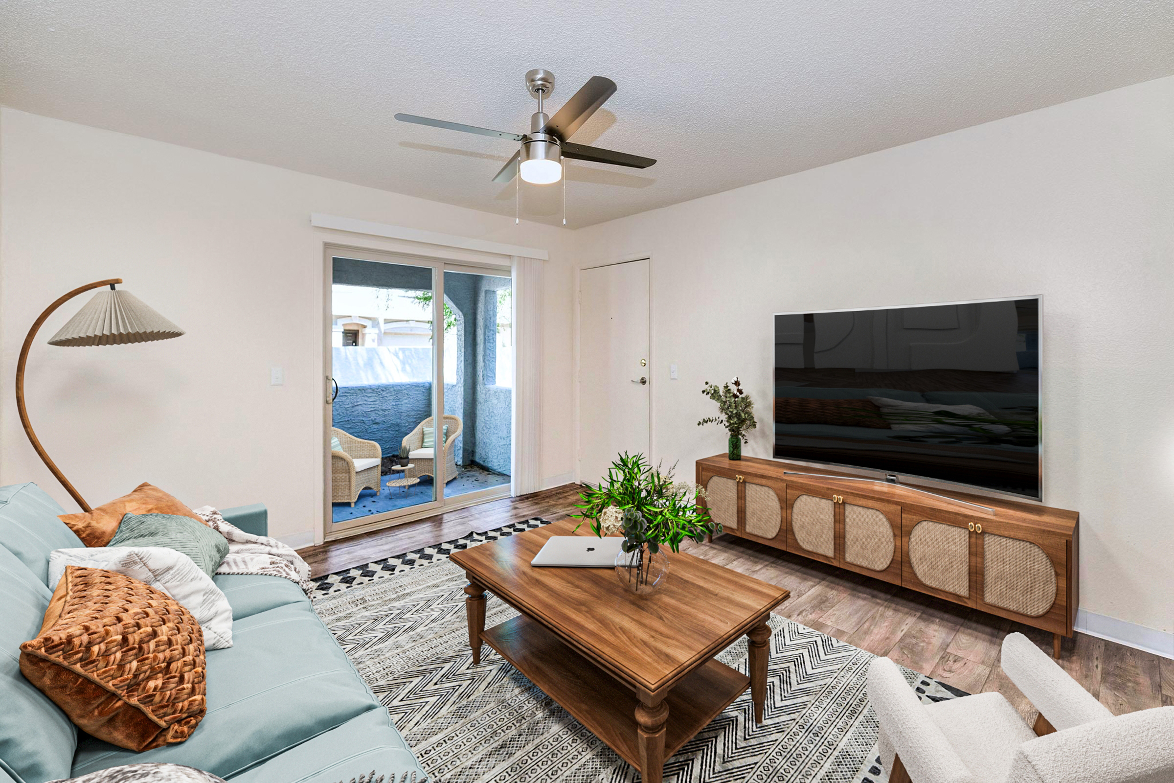A modern living room featuring a light blue sofa with decorative pillows, a wooden coffee table, and a large flat-screen TV. A ceiling fan is installed, and there is a stylish floor lamp. The room has a cozy atmosphere with a patterned area rug, and a sliding door leads to a small outdoor space.
