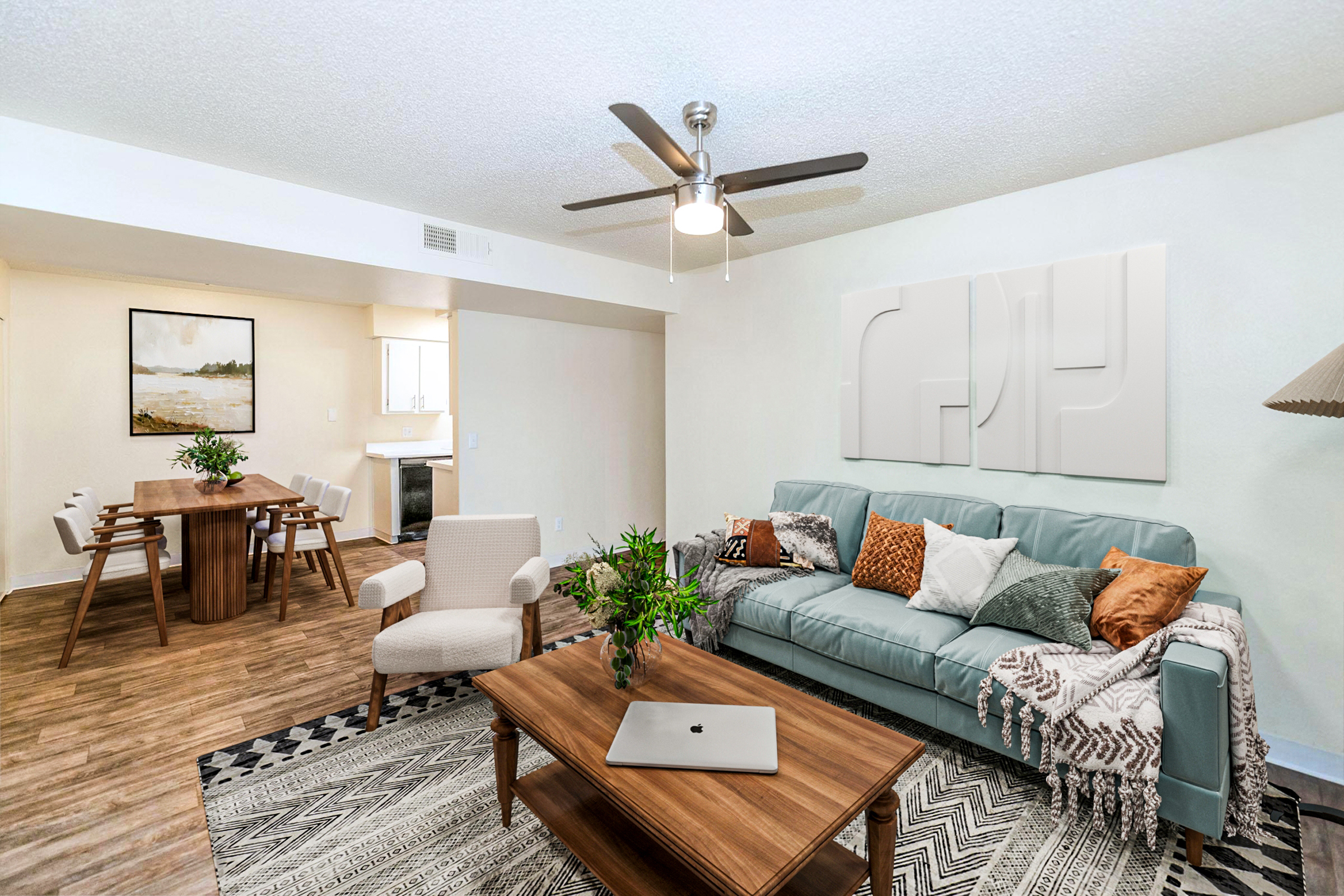A stylish living room featuring a light blue sofa adorned with decorative pillows, a wooden coffee table with a laptop on it, and a patterned rug. There’s a modern ceiling fan and a cozy armchair. In the background, a dining area with a wooden table and chairs is visible, along with a kitchen. Sunlight brightens the space.