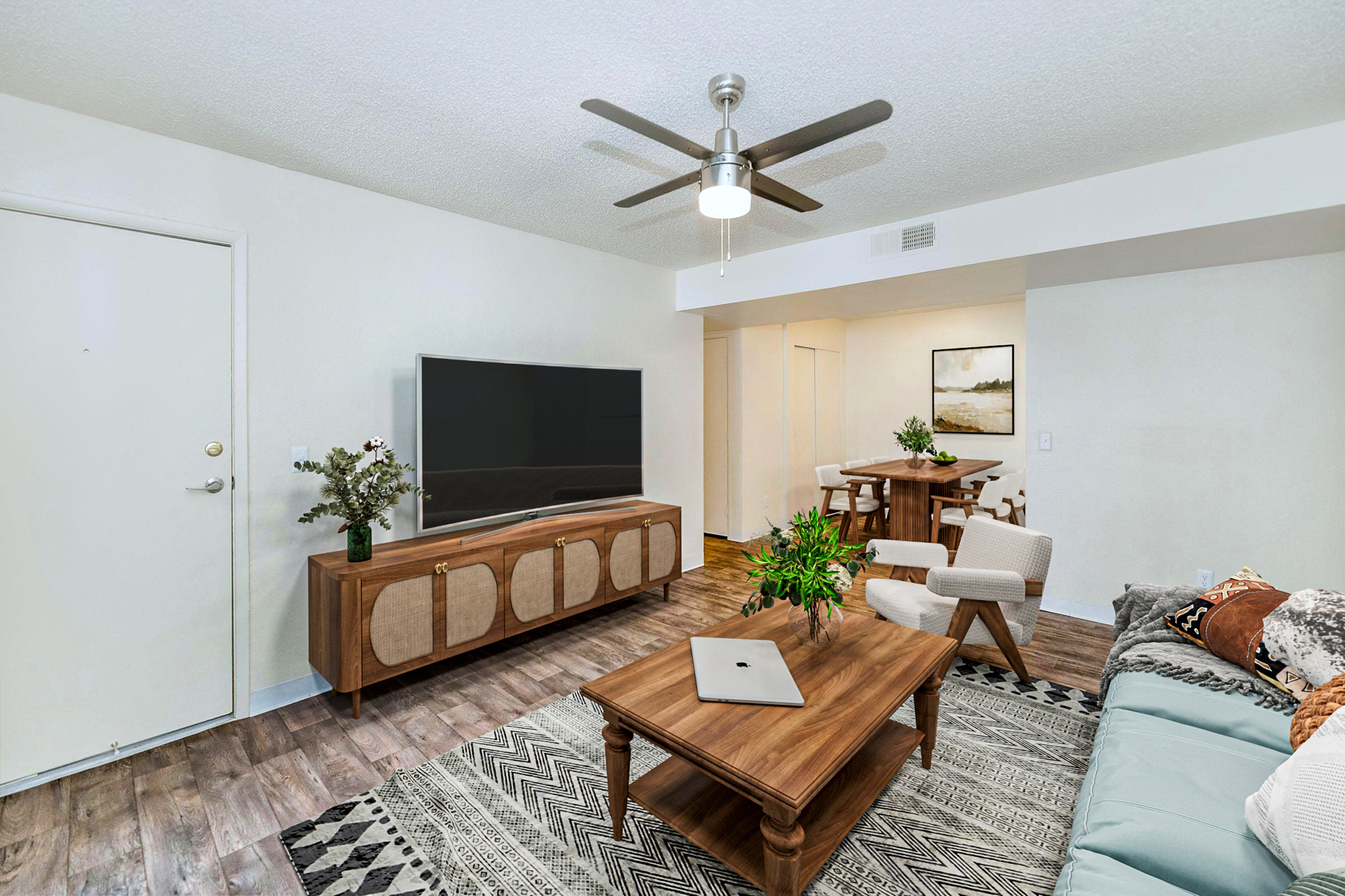 A modern living room featuring a light-colored sofa, a wooden coffee table, and a brown media console with decorative accents. A large flat-screen TV is mounted on the wall. To the side, a dining area includes a wooden table and chairs. There's a ceiling fan and decor elements like plants and artwork.