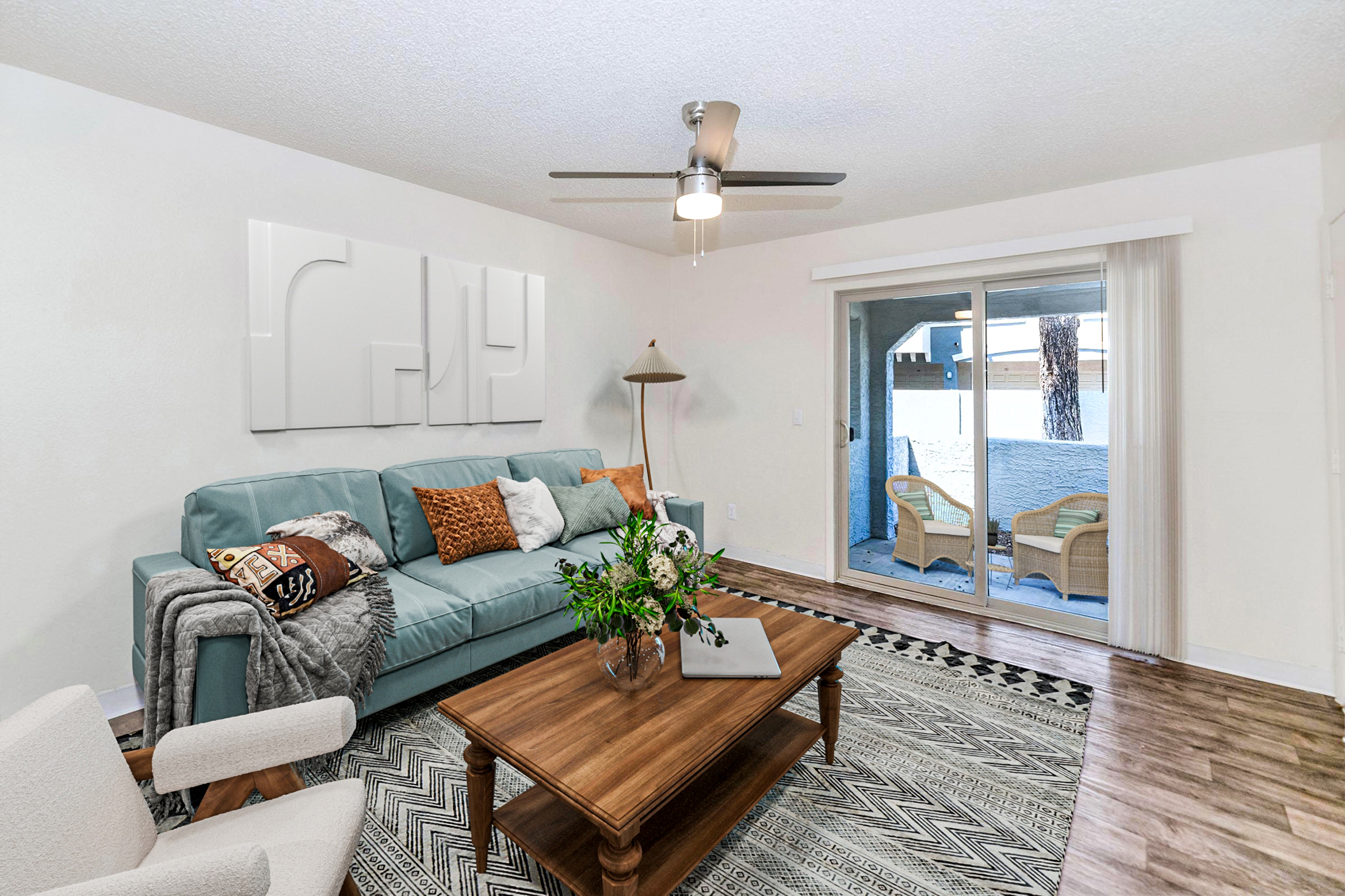 A modern living room featuring a teal sofa with decorative pillows, a wooden coffee table with a vase of flowers, and a textured area rug. There’s a minimalist wall art piece, a standing lamp, and a large glass door leading to an outdoor space. Bright and airy, with light-colored walls and wooden flooring.