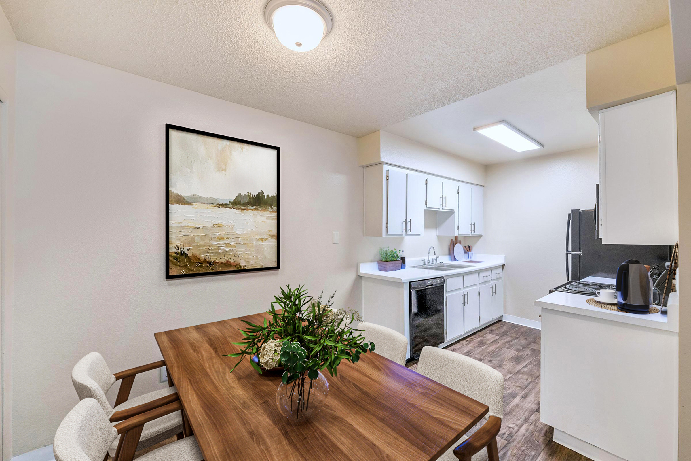 A modern kitchen and dining area featuring a wooden dining table with floral centerpieces, four cushioned chairs, and a large artwork on the wall. The kitchen is equipped with white cabinets, stainless steel appliances, and a countertop. Natural light illuminates the space, creating a welcoming atmosphere.