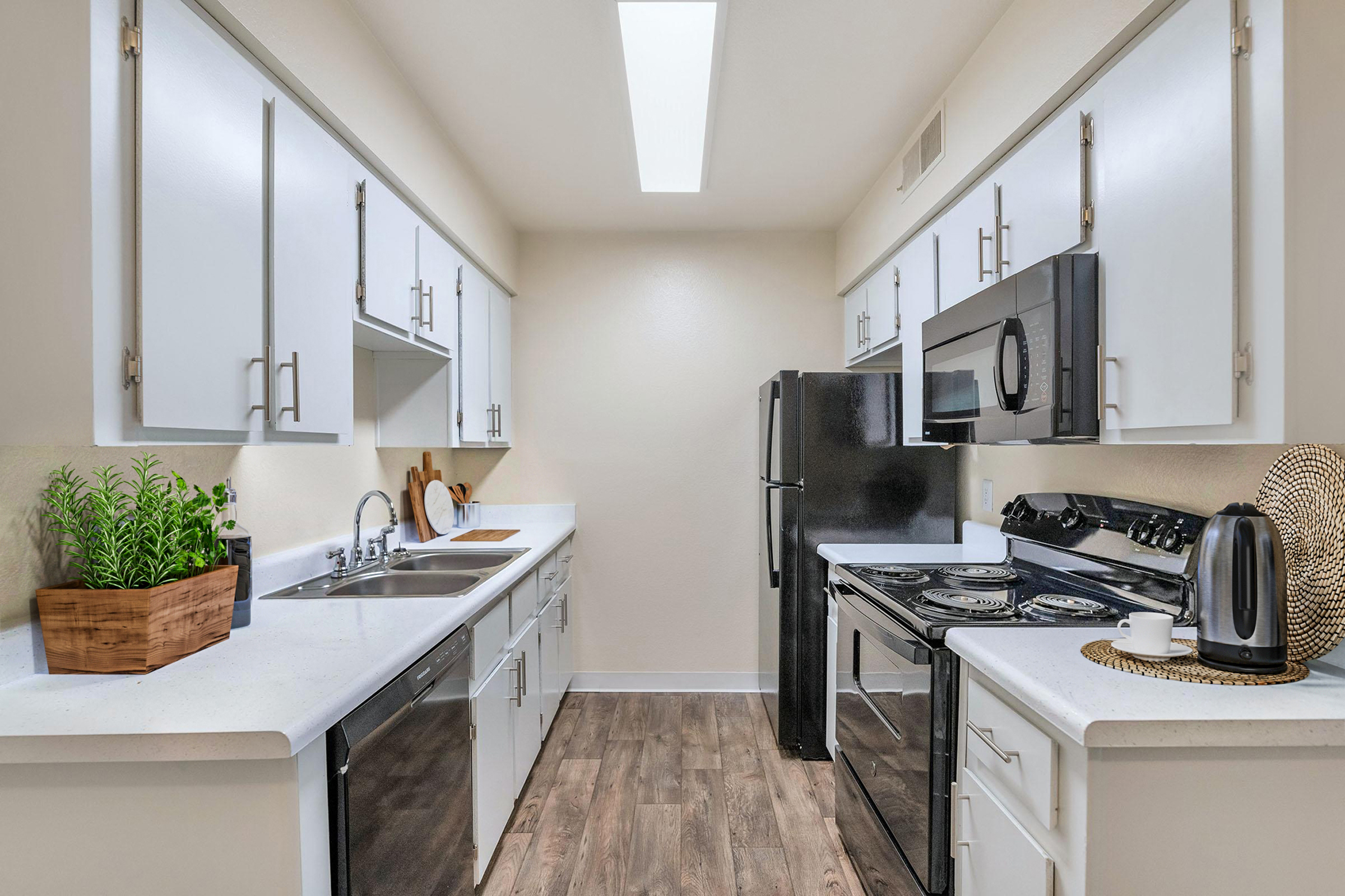 A modern kitchen featuring white cabinets, a black stove and microwave, a black refrigerator, and a sink. The countertops are light-colored, with a potted plant in a wooden container and a decorative kettle on the counter. Natural light enters from a skylight above.