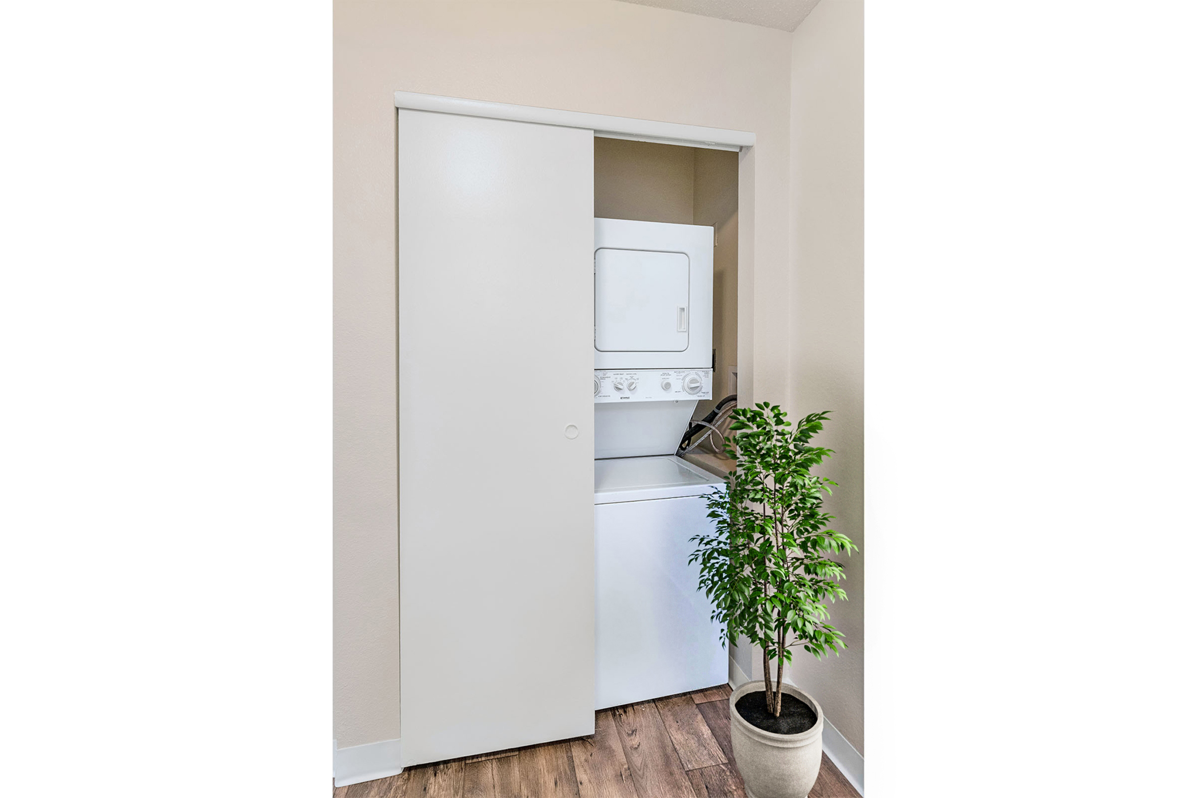 A laundry area featuring a stacked washer and dryer behind sliding white doors. A green potted plant adds a touch of decor beside the appliances, and the space has light-colored walls and wooden flooring.