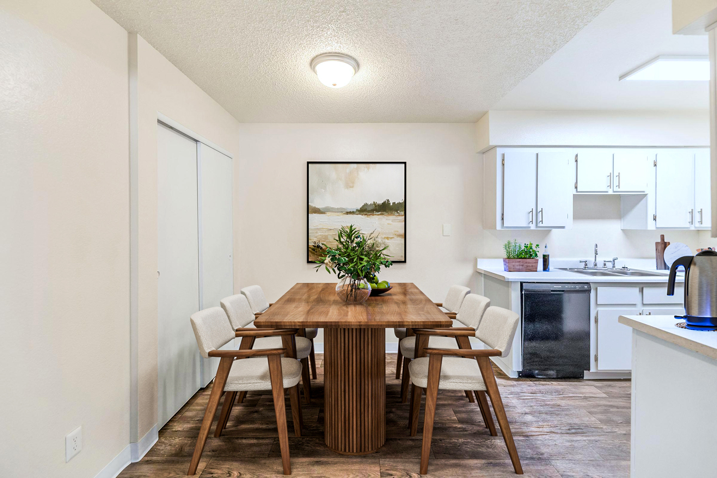 A modern dining area featuring a wooden table surrounded by light-colored chairs. There's a large artwork of a landscape on the wall, and the space has a neutral color palette. A plant is placed in the center of the table, and a kitchen area with white cabinetry and appliances is visible in the background.