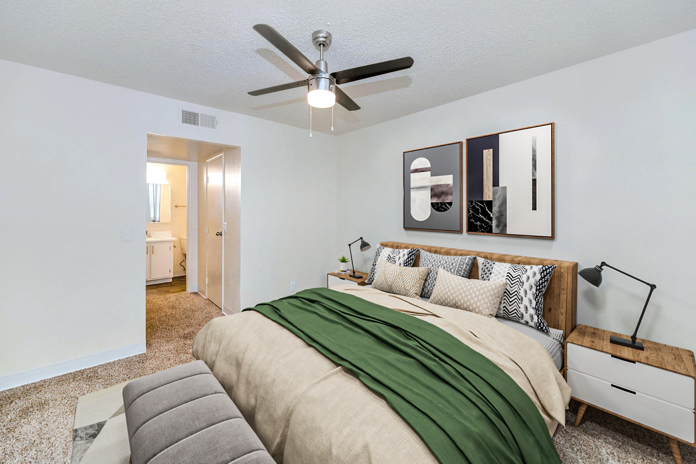 A modern bedroom featuring a large bed with a green and beige throw blanket, two decorative pillows, and stylish wall art. There is a ceiling fan, a bedside table with a lamp, and a bench at the foot of the bed. A doorway leads to another room, visible in the background. The decor is bright and inviting.