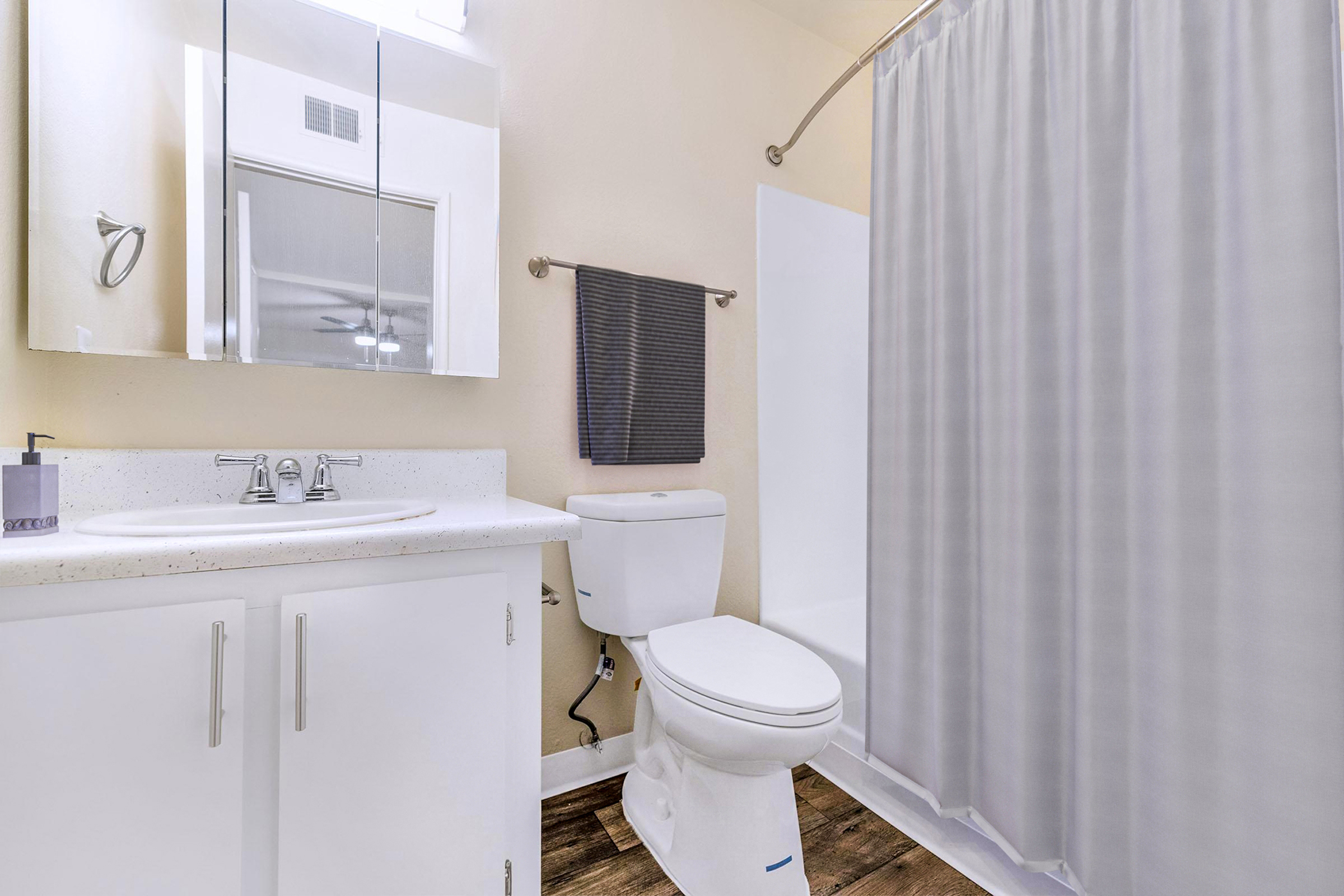 A clean, modern bathroom featuring a white countertop sink, a toilet, and a bathtub with a shower curtain. The walls are painted a light color, and there is a wall mirror and a dark towel hanging. The flooring is wooden, contributing to a contemporary aesthetic.