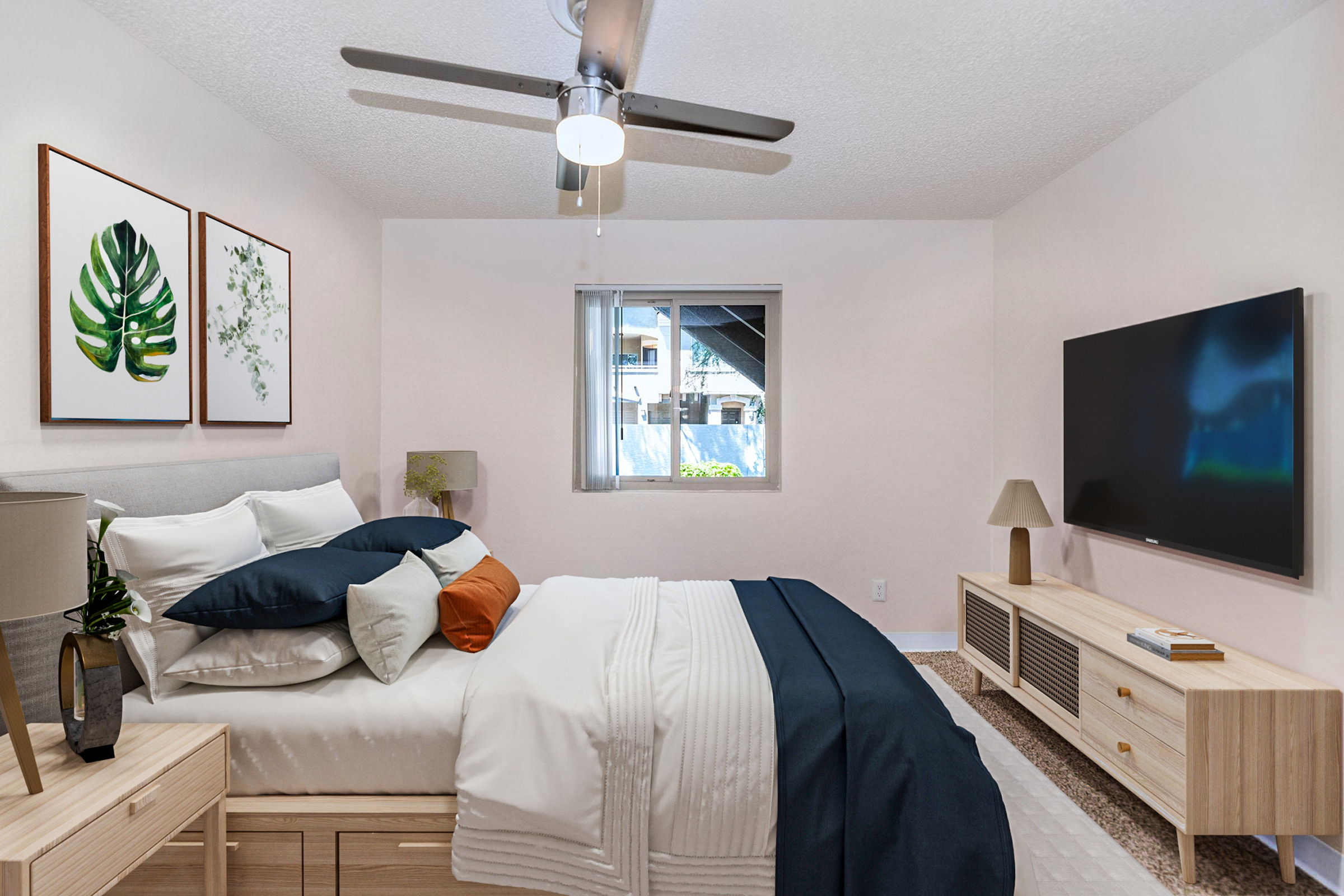 A cozy bedroom featuring a light color scheme with a neatly made bed adorned with decorative pillows. Two framed botanical prints are on the wall, and a modern ceiling fan is above. A television is mounted on the wall opposite the bed, accompanied by a stylish TV stand. Natural light filters through the window.
