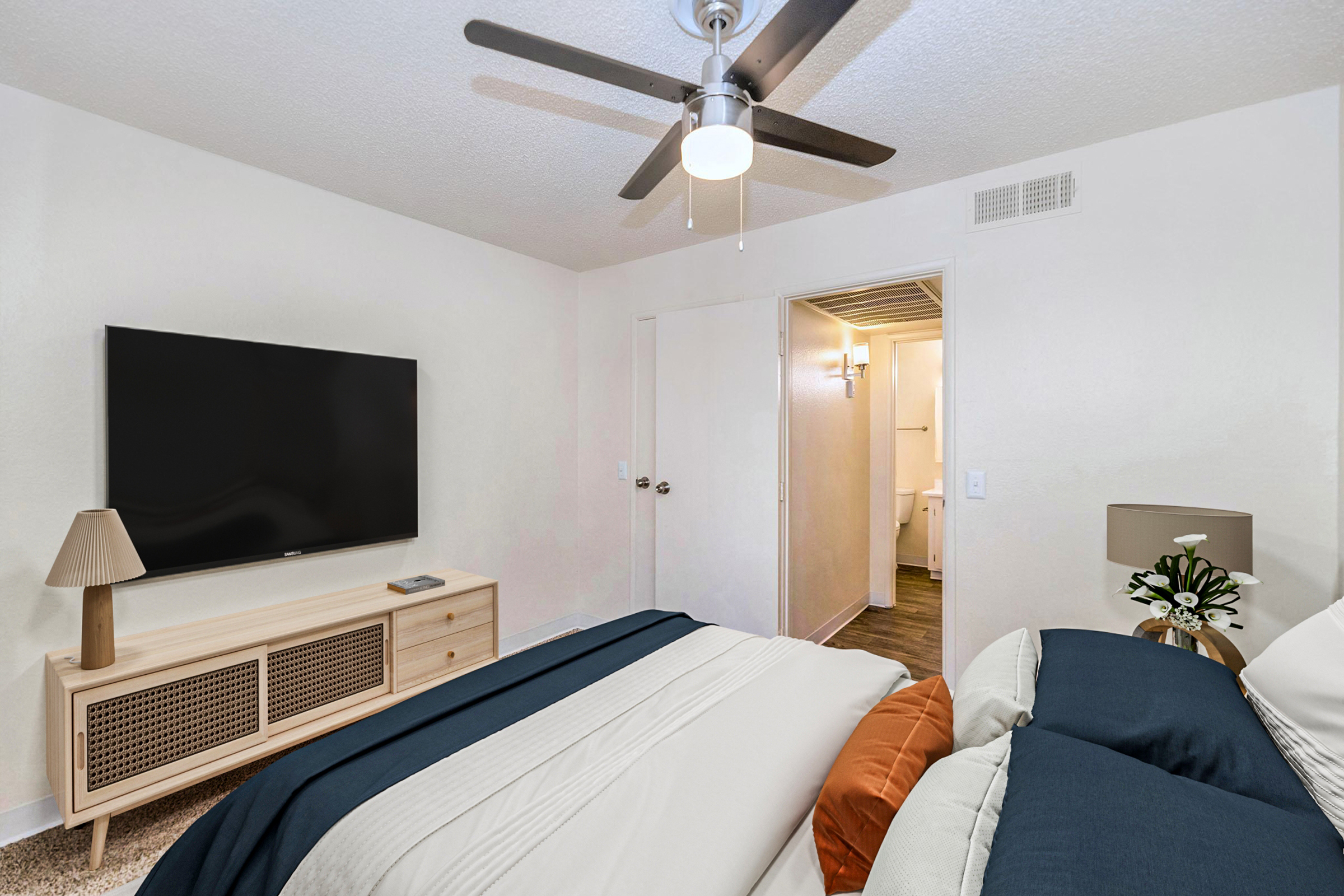 A cozy bedroom featuring a bed with navy and cream bedding, a light wooden dresser with a television on top, a lamp, and a plant. A ceiling fan is visible, and a doorway leads to a bathroom area. The walls are painted white, creating a clean and inviting atmosphere.