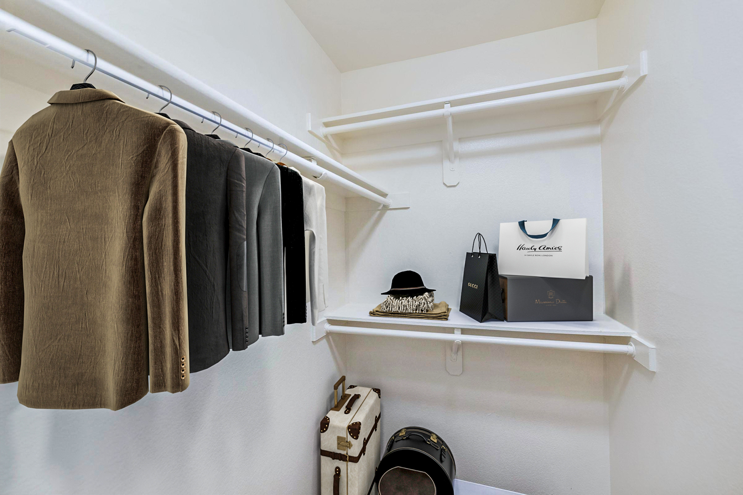 A neatly organized closet featuring a row of suits on hangers, a black hat, a folded item, and two stylish bags. The space is illuminated, showcasing a clean and minimalistic design with white walls and shelving.