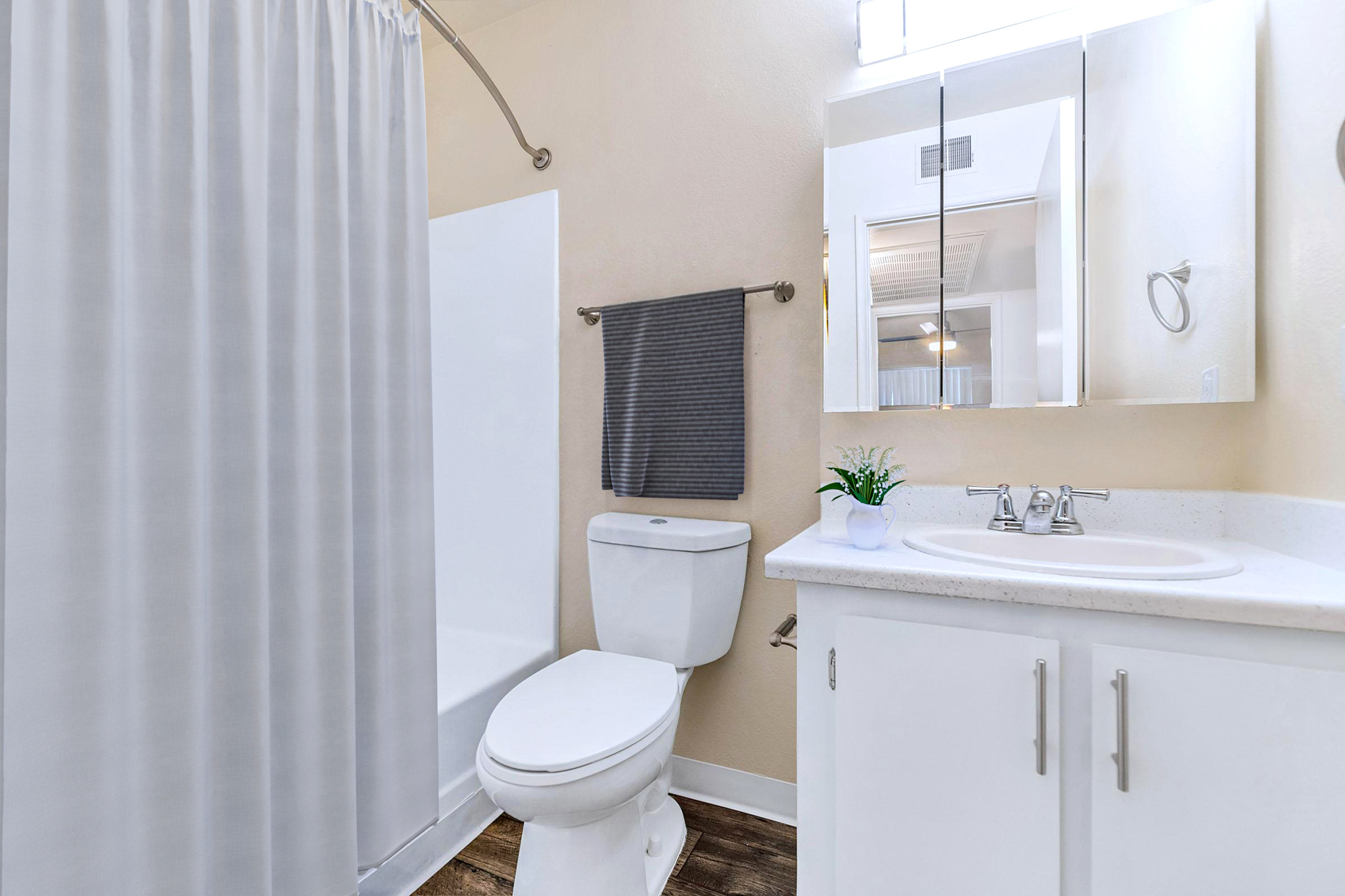 A clean and modern bathroom featuring a white bathtub with a shower curtain, a white toilet, a sink with a countertop, and a mirror. The walls are light-colored, and there’s a small plant on the sink. A gray towel is hung nearby, adding a touch of decor to the space.