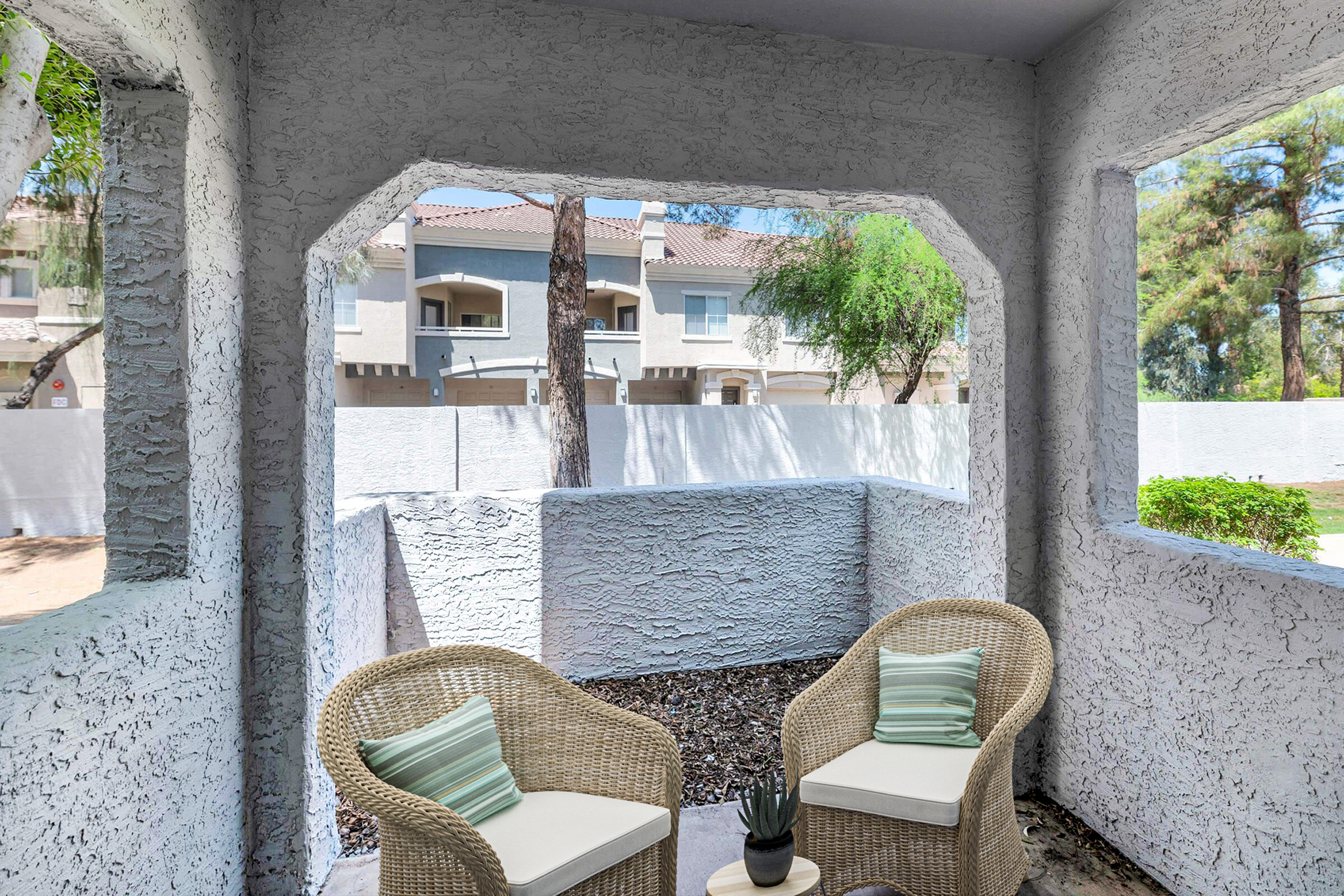 A cozy outdoor patio with two woven chairs and decorative cushions, surrounded by textured white walls. The view features a landscaped area with trees and neighboring buildings in the background, creating a serene and inviting atmosphere.