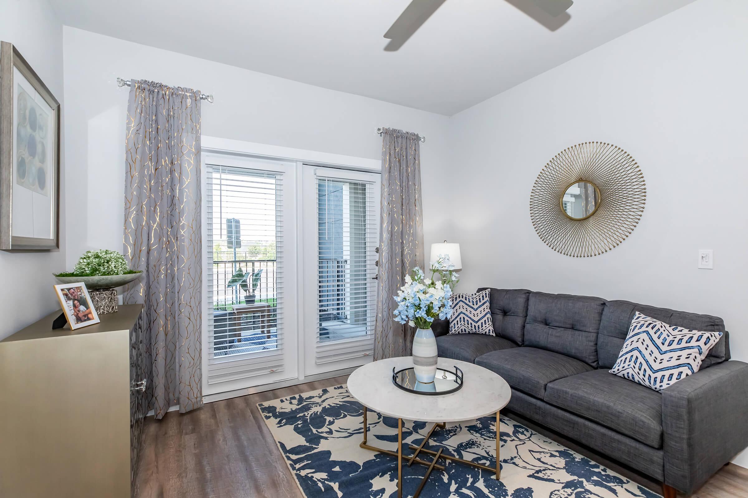 A modern living room featuring a gray sofa with decorative pillows, a glass coffee table on a patterned rug, and a stylish wall mirror. Sheer curtains drape over sliding glass doors leading to a balcony, with a small potted plant and a framed photo on a sleek side table, creating a cozy and inviting atmosphere.