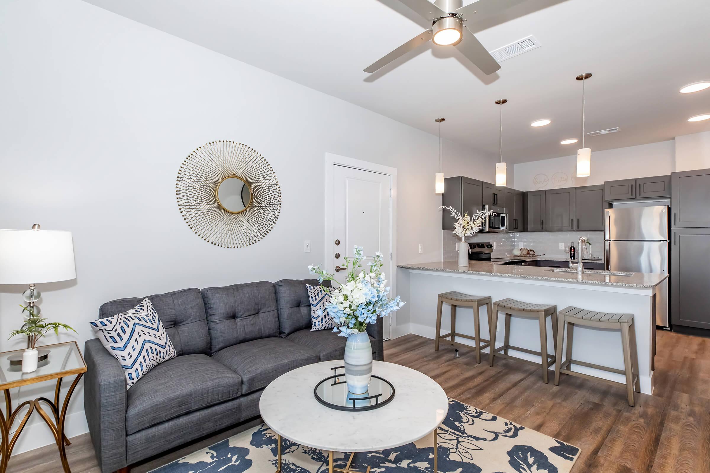 A modern living room featuring a gray sofa with patterned cushions, a round coffee table, and a decorative plant. In the background, there is a shiny kitchen with stainless steel appliances and bar stools. A stylish mirror and pendant lights enhance the contemporary aesthetic.