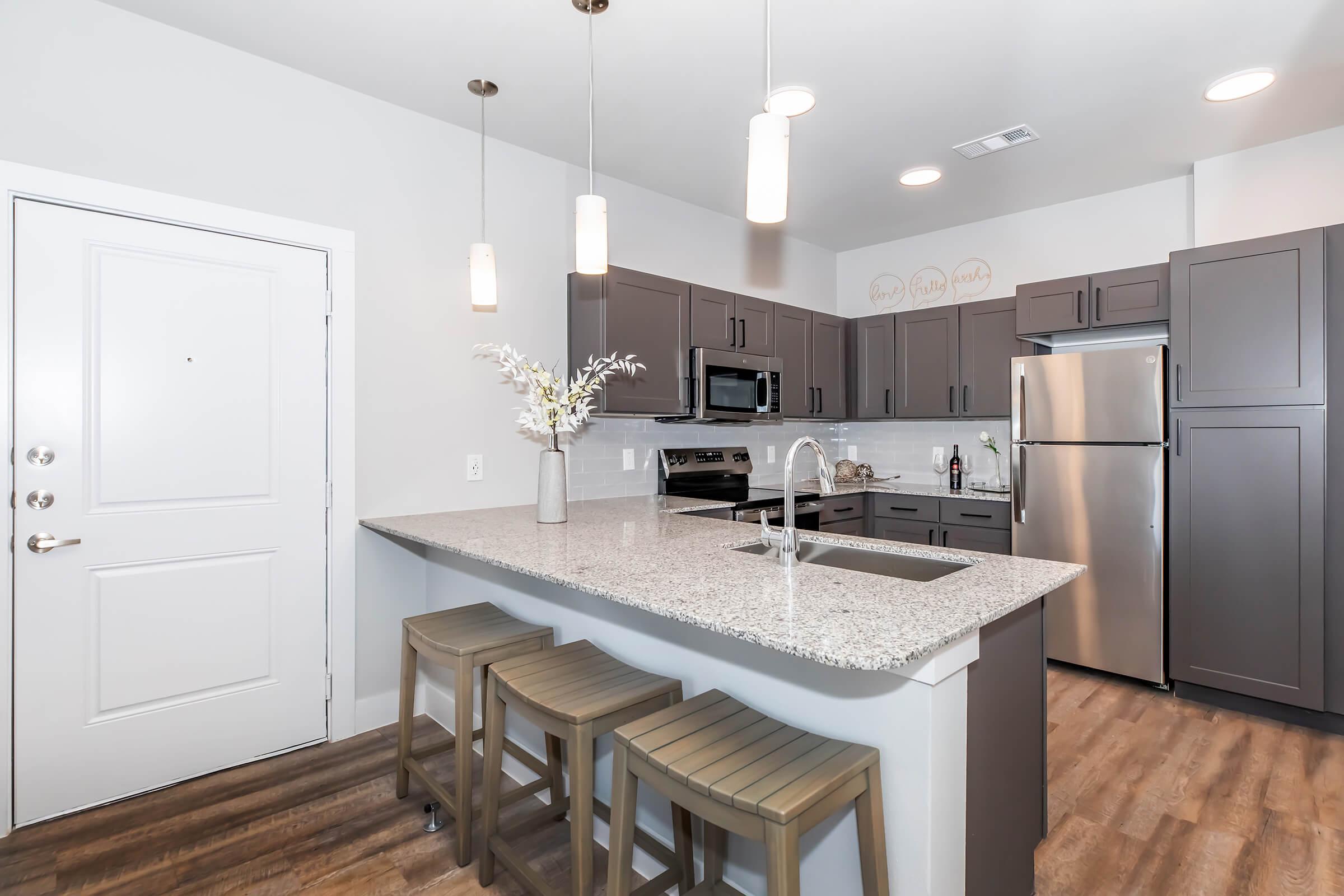 Modern kitchen with gray cabinets, stainless steel appliances, and a large granite island with three wooden stools. The space features bright pendant lighting and a spacious layout. A doorway leads to the entrance, while decorative items adorn the counter, enhancing the inviting atmosphere.
