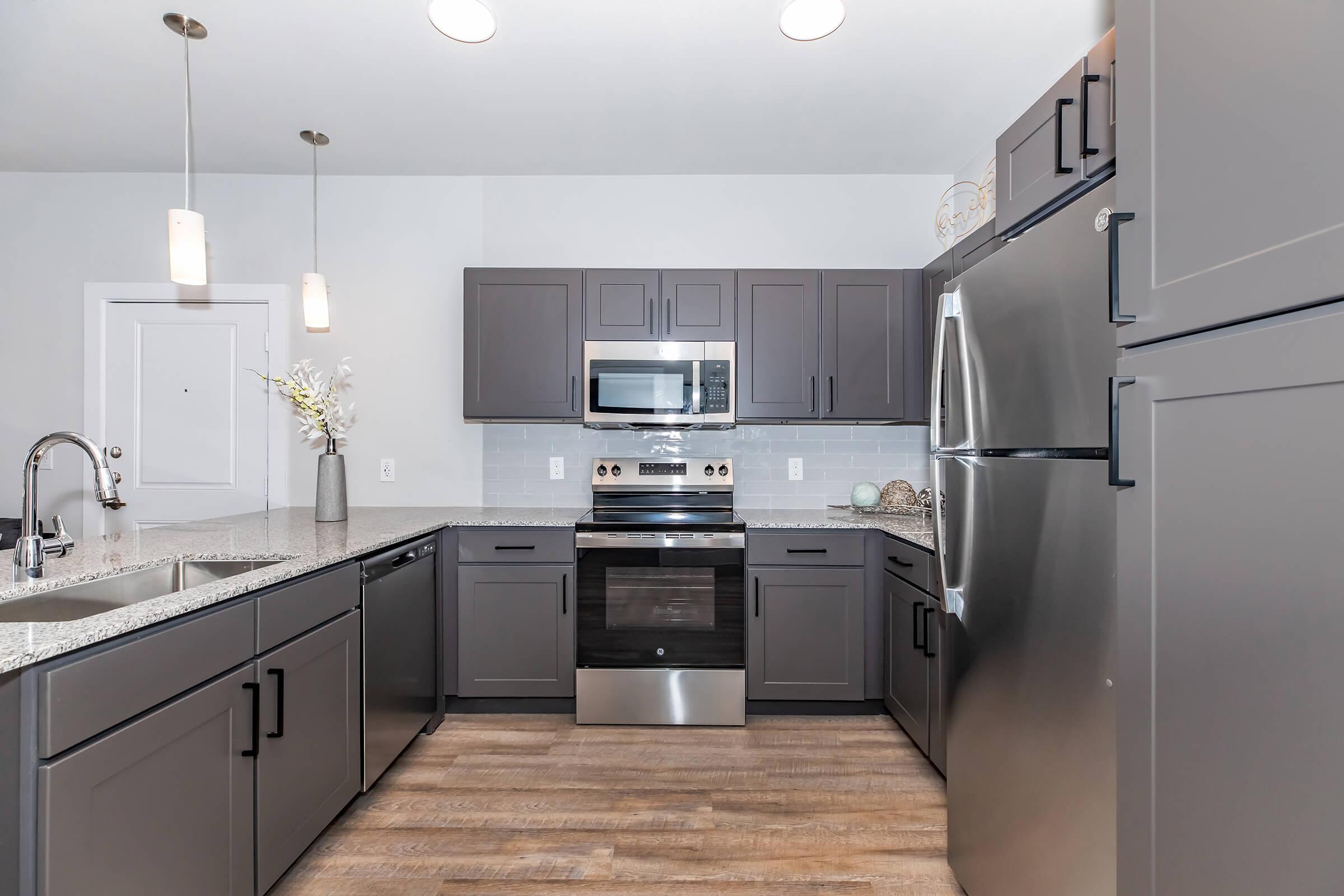 Modern kitchen featuring dark gray cabinetry, stainless steel appliances, a sleek microwave, and a granite countertop. The open layout is complemented by pendant lighting and wood-like flooring, creating a contemporary and inviting atmosphere.