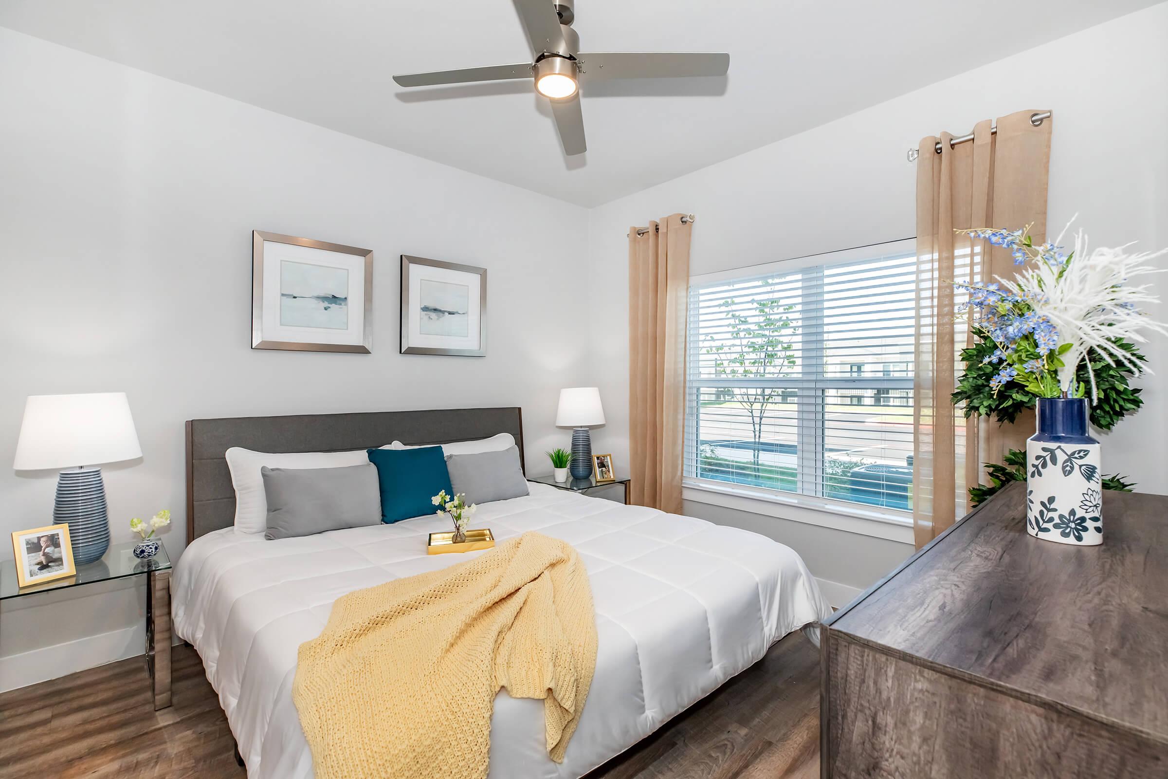 A modern and inviting bedroom featuring a queen-sized bed with gray and white bedding, a light yellow throw blanket, and decorative pillows. Two bedside tables with lamps and framed artwork adorn the space. A large window with sheer curtains lets in natural light, complemented by potted plants and stylish decor.