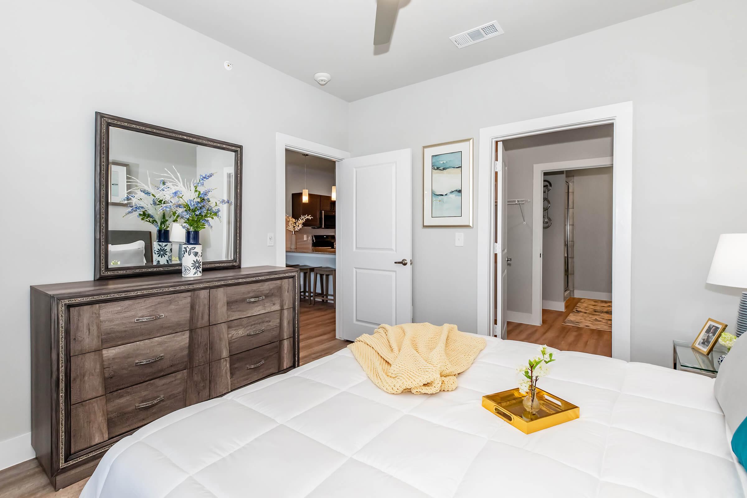 A modern bedroom featuring a neatly made bed with a white comforter, a decorative yellow blanket draped over a nightstand, and a stylish dresser with a mirror. Fresh flowers in a vase and framed artwork add a touch of elegance. A doorway leads to another room, enhancing the spacious feel.