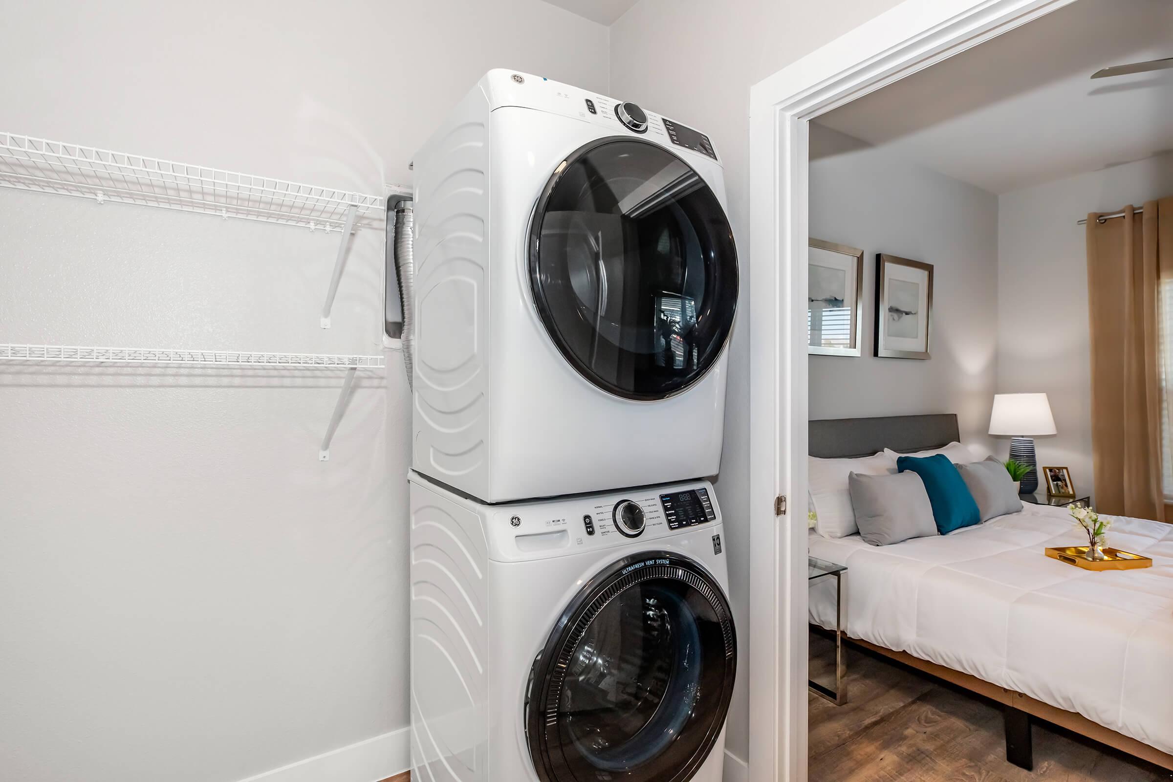 Stacked washer and dryer units in a bright laundry nook, with a glimpse of a cozy bedroom featuring a bed with white linens and accent pillows, and a small table with flowers. A doorframe leads into the adjacent room, enhancing the inviting atmosphere of the space.